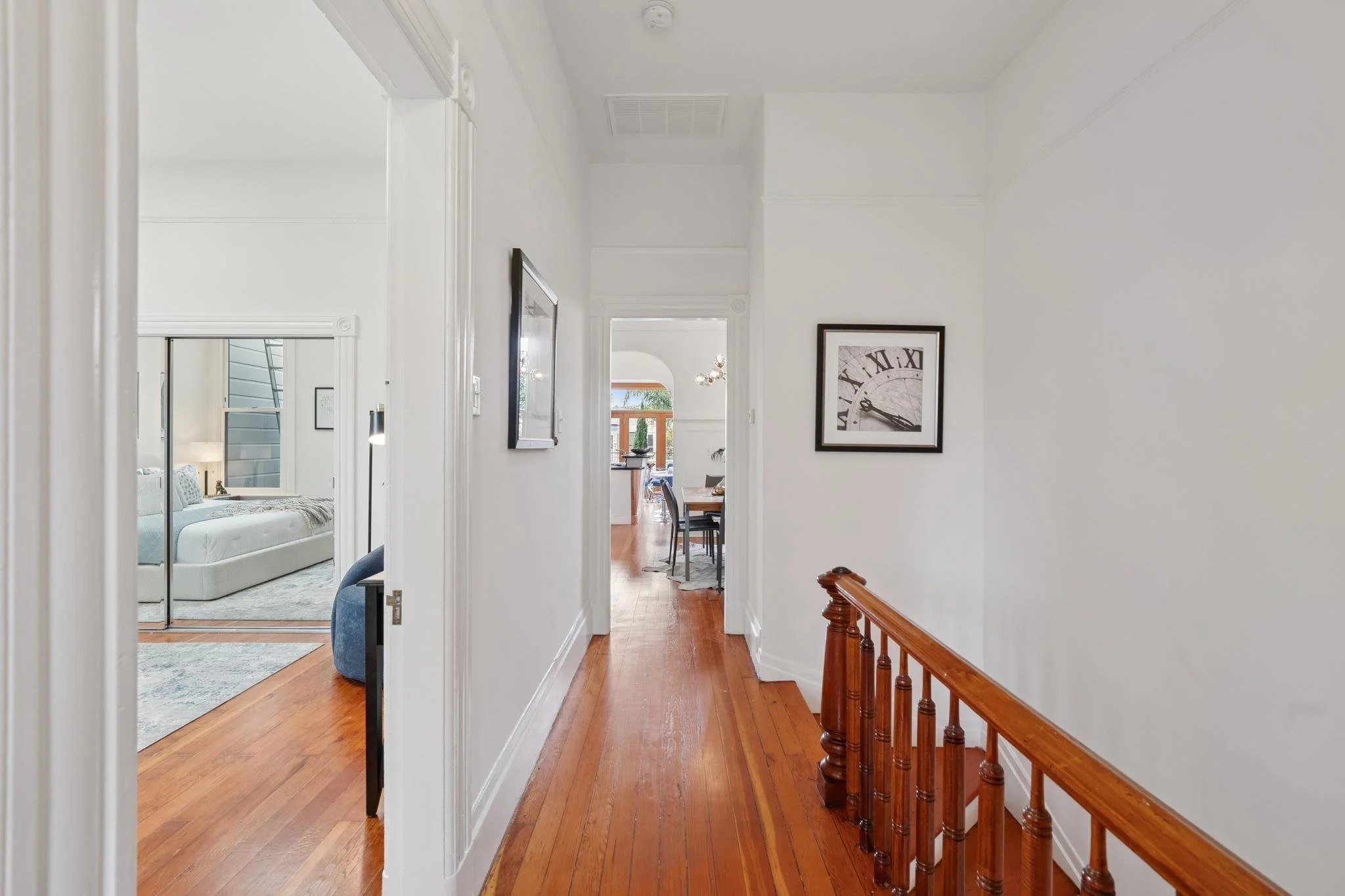 Hallway with white walls and wooden floor, leading to dining and living areas with visible furniture, including a bed in a bedroom on the left.