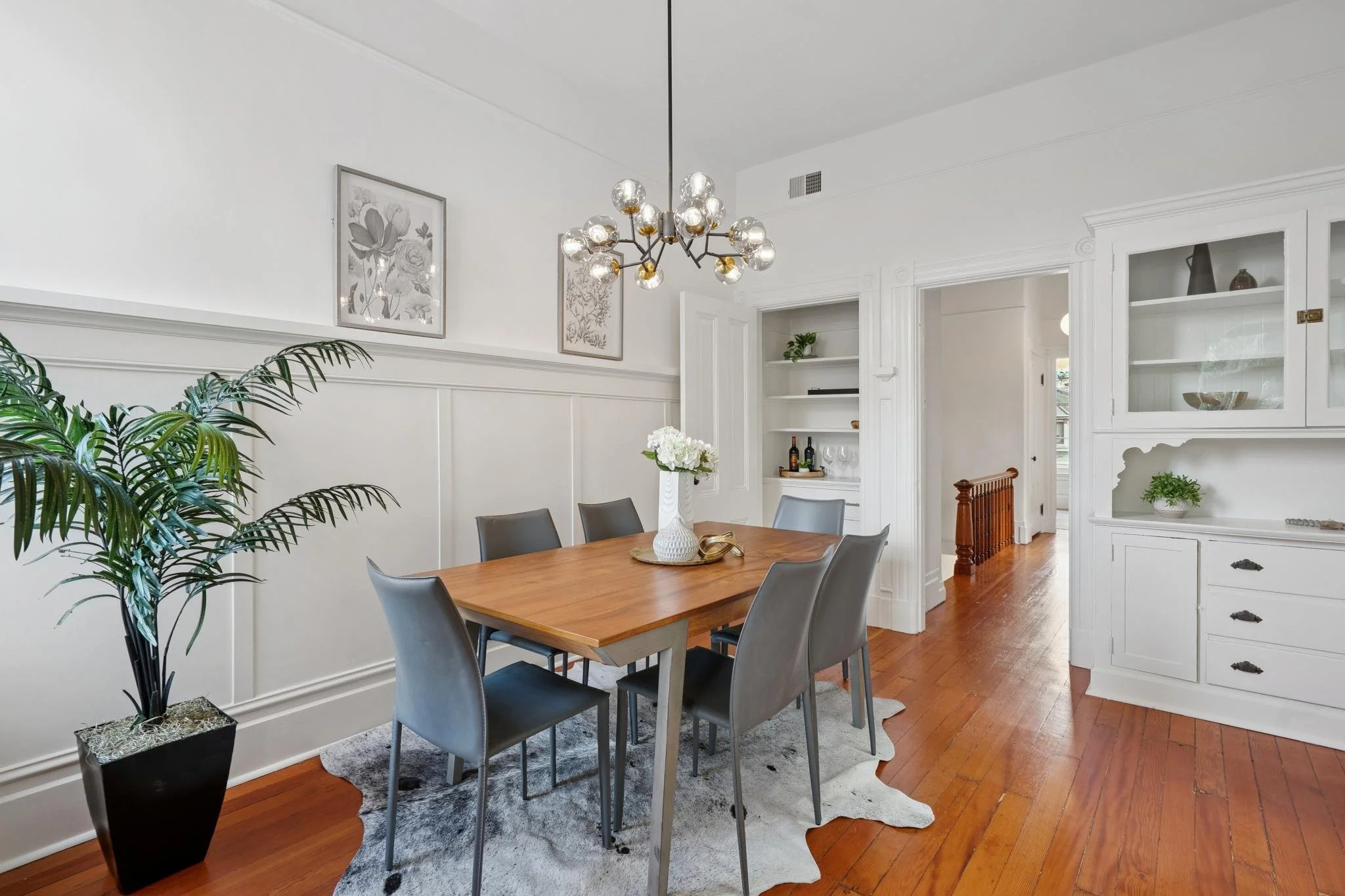 A dining room with a wooden table, six gray chairs, a large potted plant, white built-in shelves, framed botanical art, and a modern chandelier.