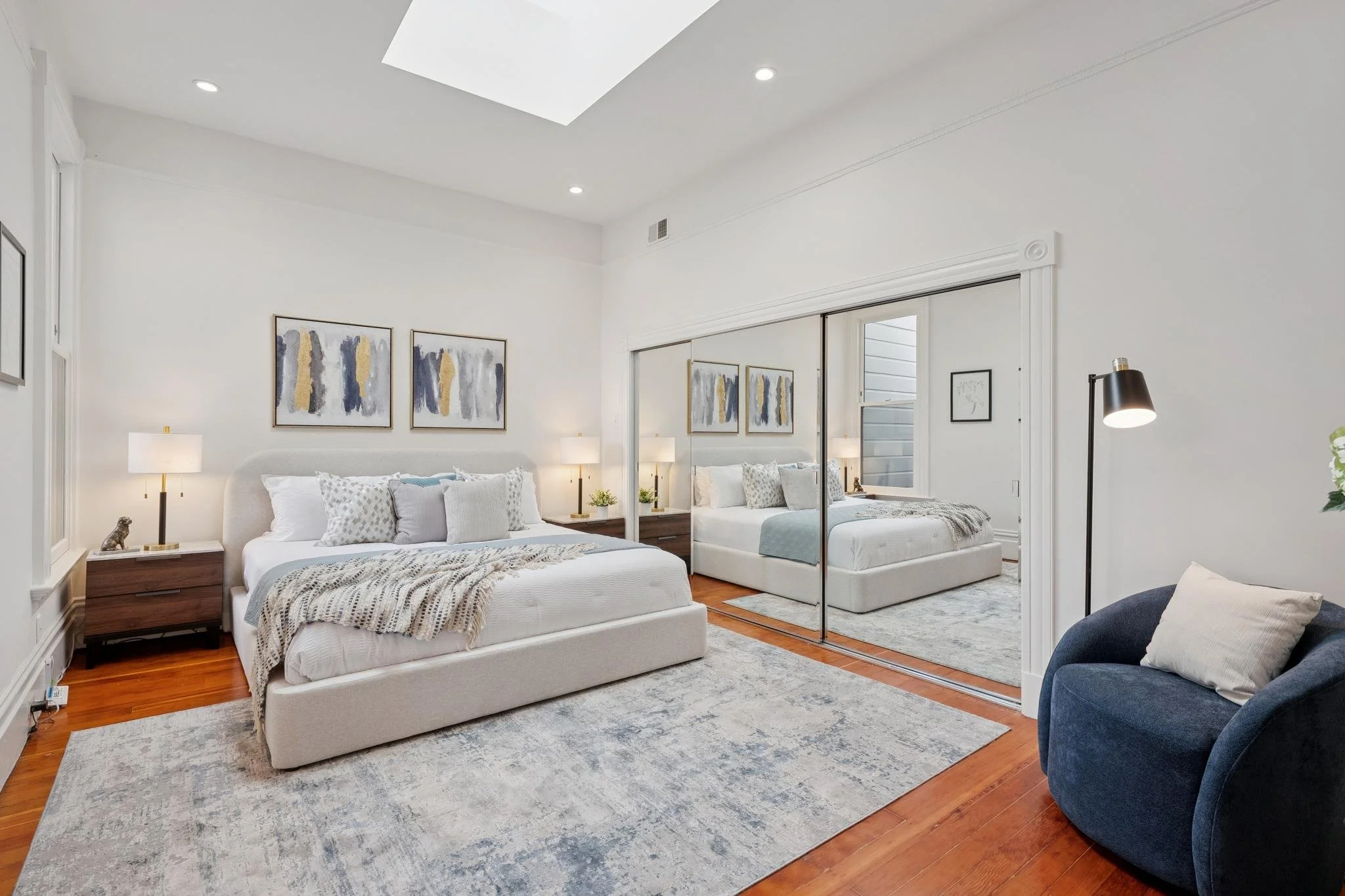 A modern bedroom with a large bed, nightstands, bedside lamps, abstract artwork, mirrored closet doors, and a blue accent chair, all with a wooden floor and white walls.