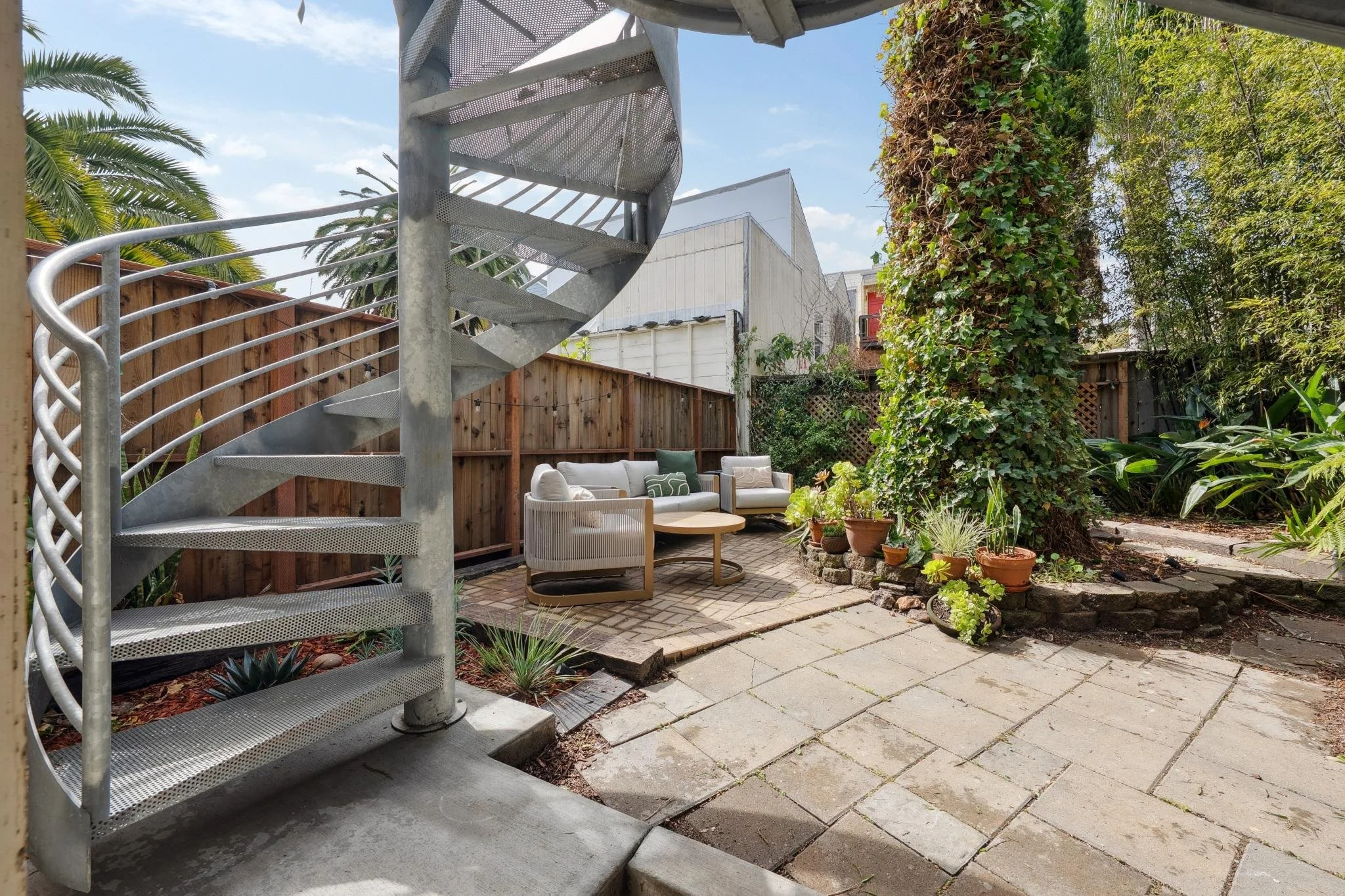Backyard patio with spiral metal staircase, outdoor seating with sofas and a coffee table, surrounded by potted plants, trees, and a wooden fence, on a sunny day.