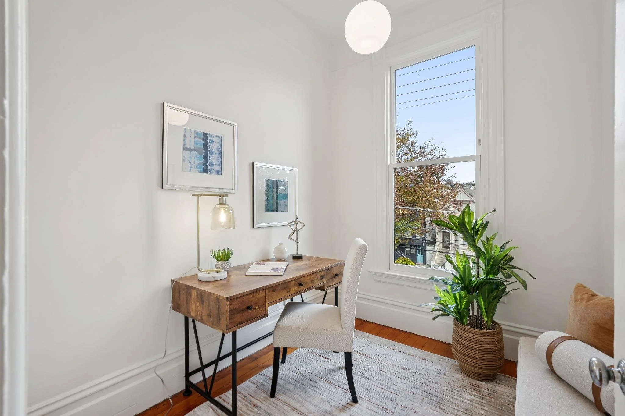 A bright home office with a wooden desk, white upholstered chair, framed artwork, a small plant, a decorative lamp, and a large window with a view of trees and rooftops, sunlight streaming in.