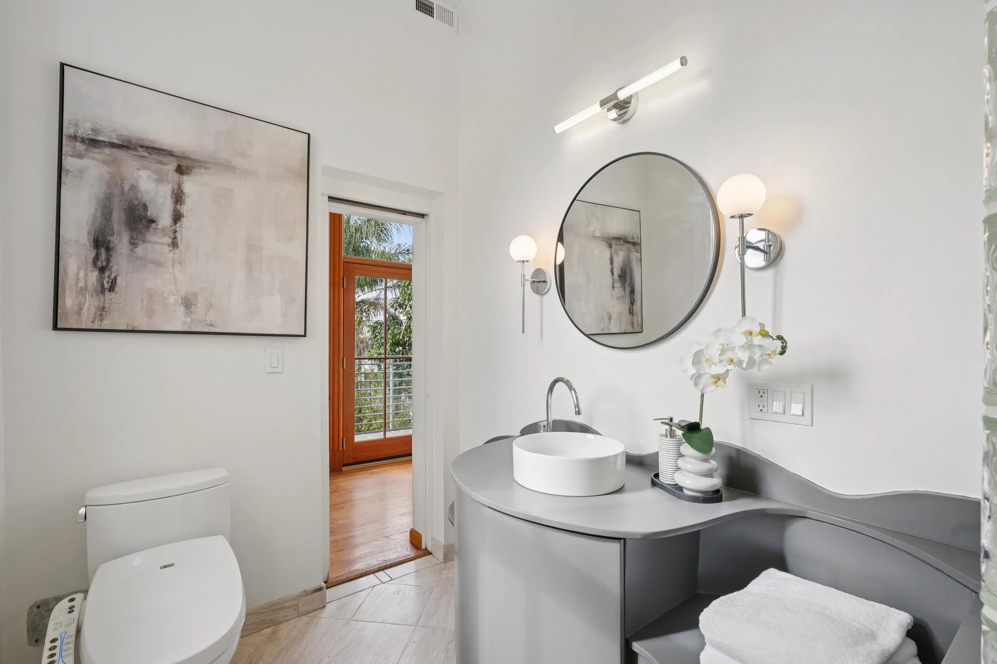 Modern bathroom with white walls, a large round mirror, wall-mounted sconces with globe lights, gray vanity with a vessel sink, a white orchid, and artwork on the wall. A door leads to a balcony with a wooden railing.