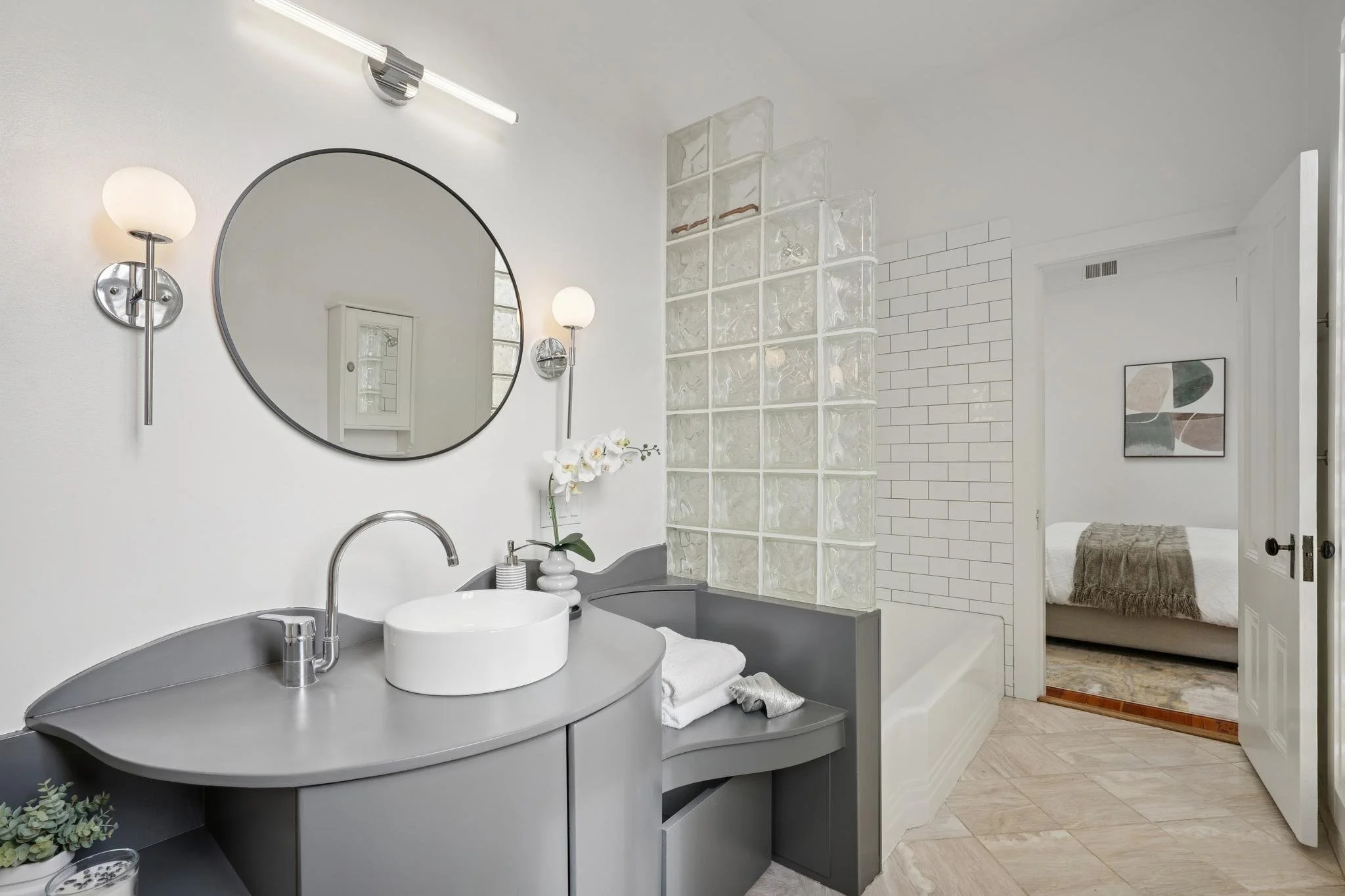 Modern bathroom with a round mirror, wall-mounted lights, a gray vanity with a white vessel sink, a glass block wall, and a connected bathtub. A view into a bedroom with a bed, artwork, and a blanket is visible through an open door.