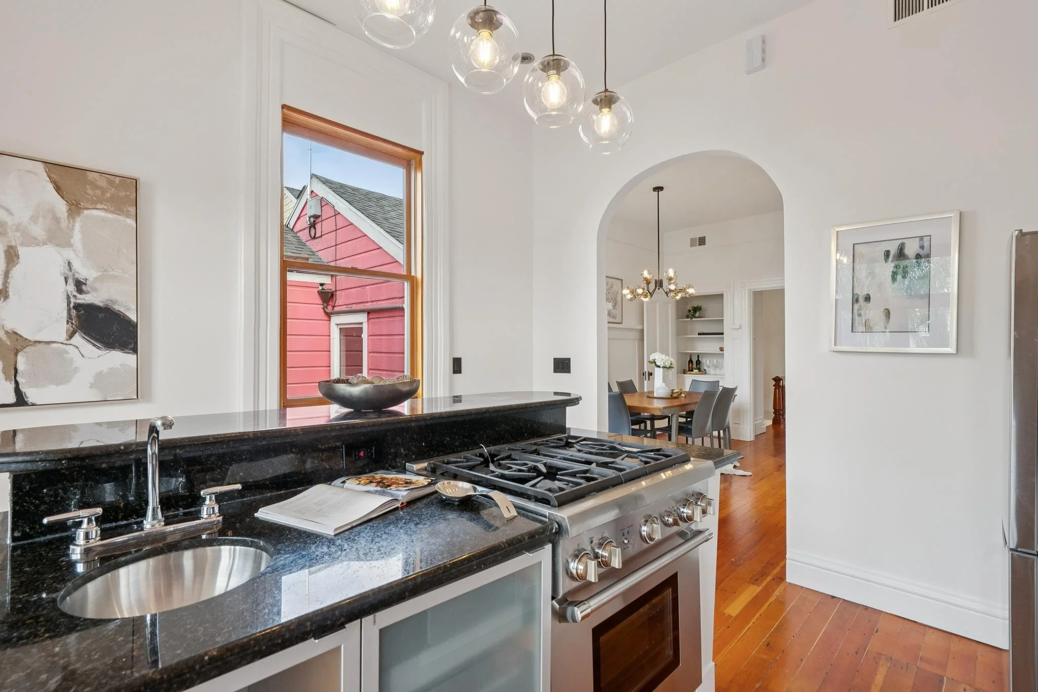 Kitchen with black granite countertops, stainless steel stove, a small circular sink, a cookbook open on the counter, and a window showing a red house outside. The room leads into a dining area with a wooden table and gray chairs, hanging light fixtu