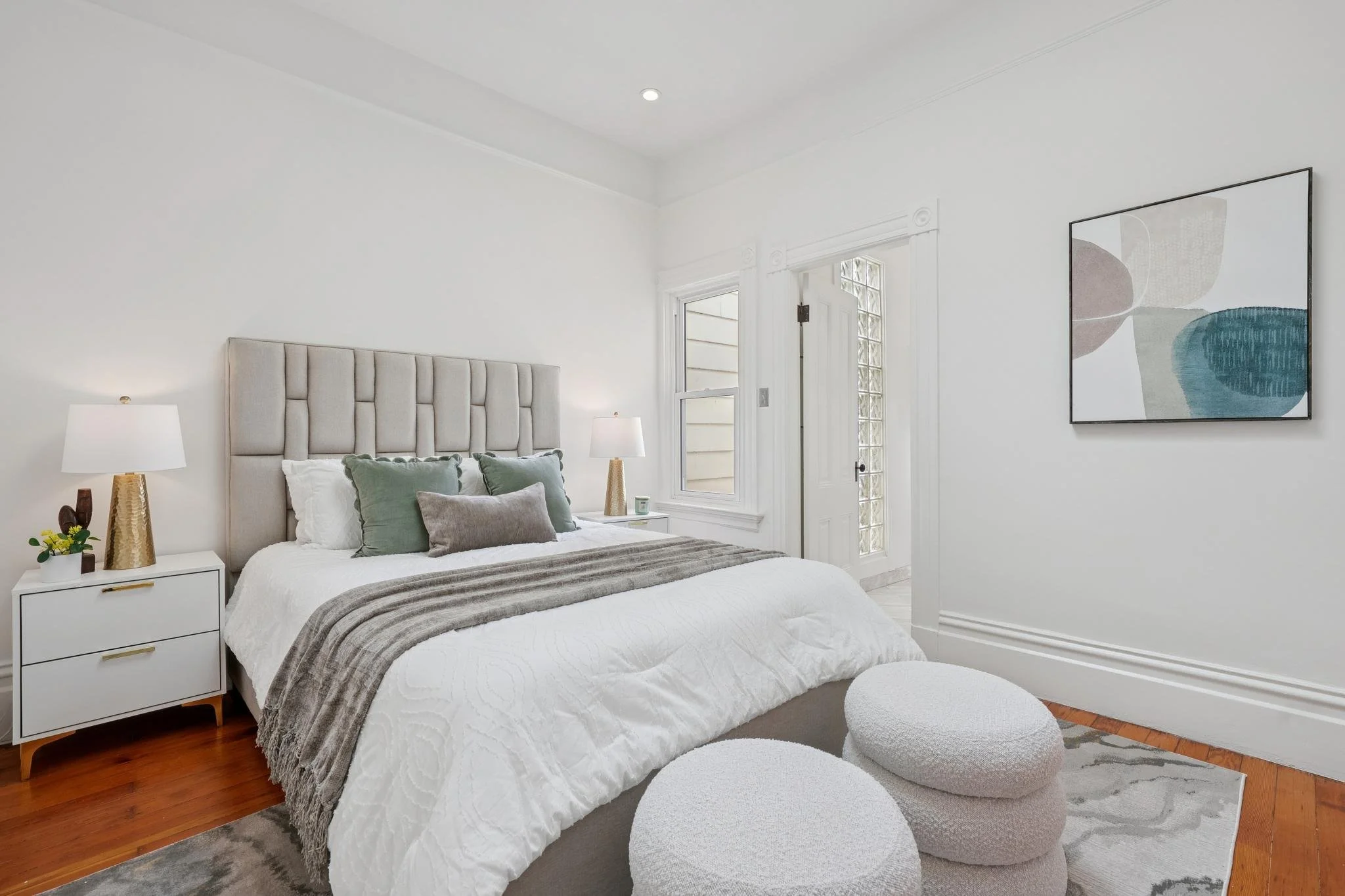 A bedroom with a bed, nightstands with lamps, and artwork on the wall. The room has white walls, a window, hardwood floors, and minimalist decor.