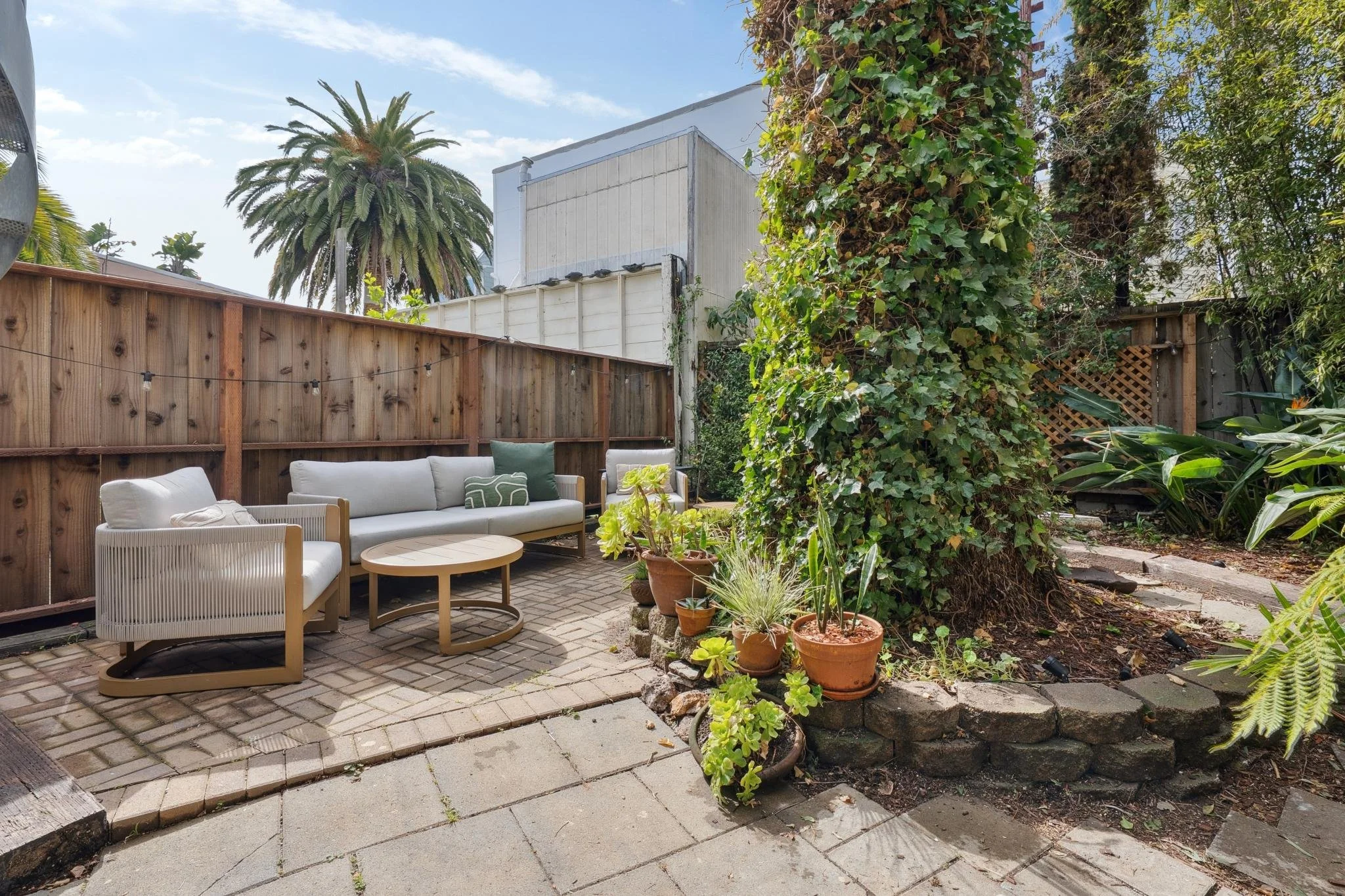 Outdoor backyard patio with white cushioned furniture, potted plants, and a tall ivy-covered tree, surrounded by a wooden fence and lush greenery.