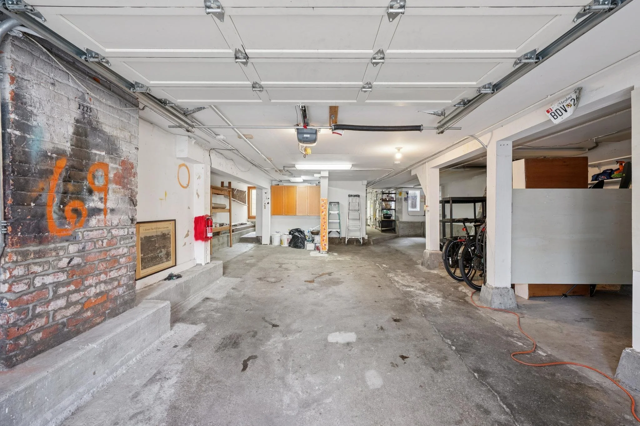 Interior of a spacious two-car garage with a concrete floor, white walls, and exposed garage door mechanics overhead. Various tools, bikes, a ladder, wooden shelves, and cabinets are scattered throughout.