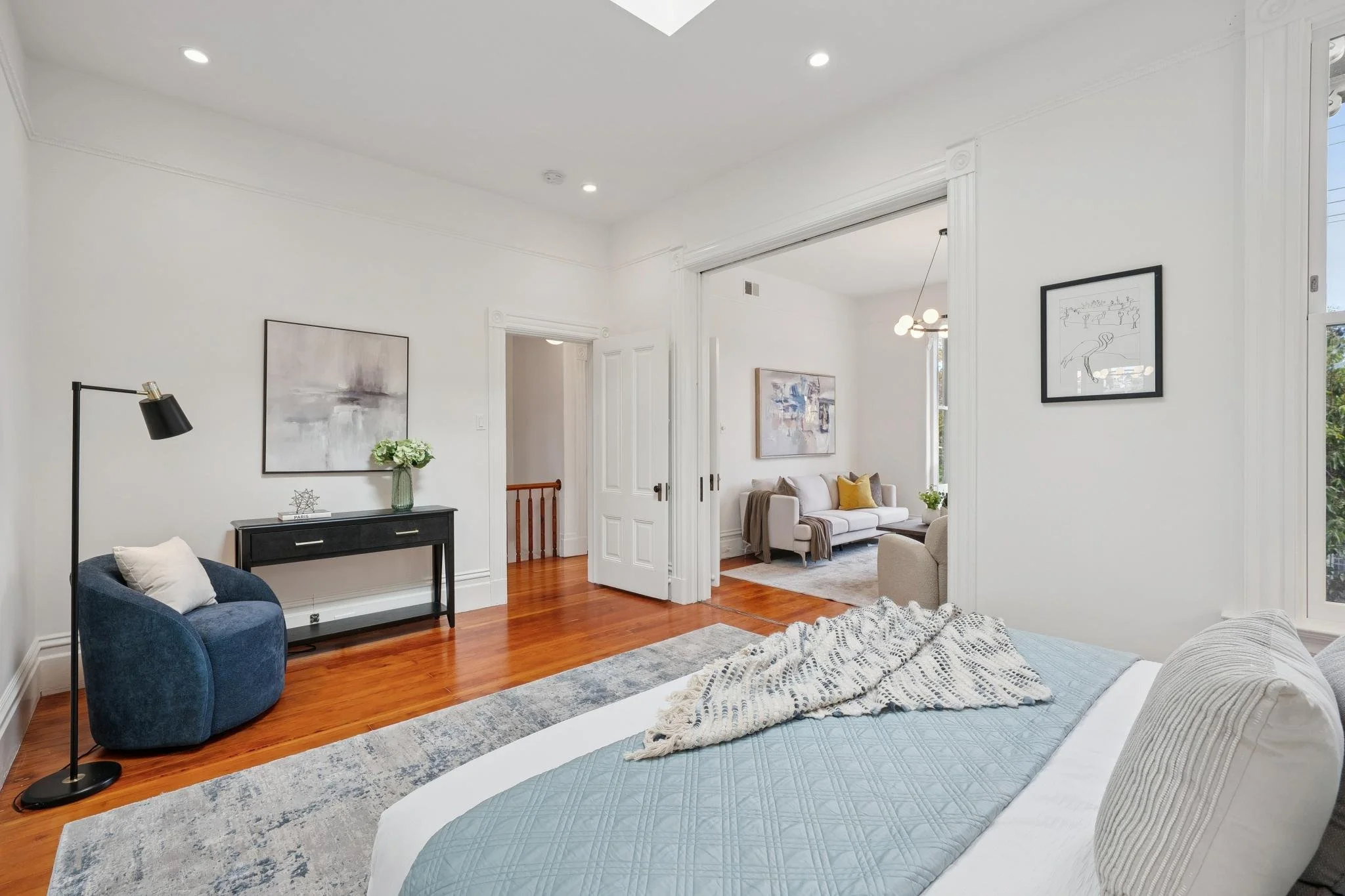 A bright bedroom with a white bed, blue quilt, and throw blanket. To the left, a blue armchair with pillow and black floor lamp. A black console with a flower arrangement and decorative object. Past a doorway, a living room with a beige sofa, pillows