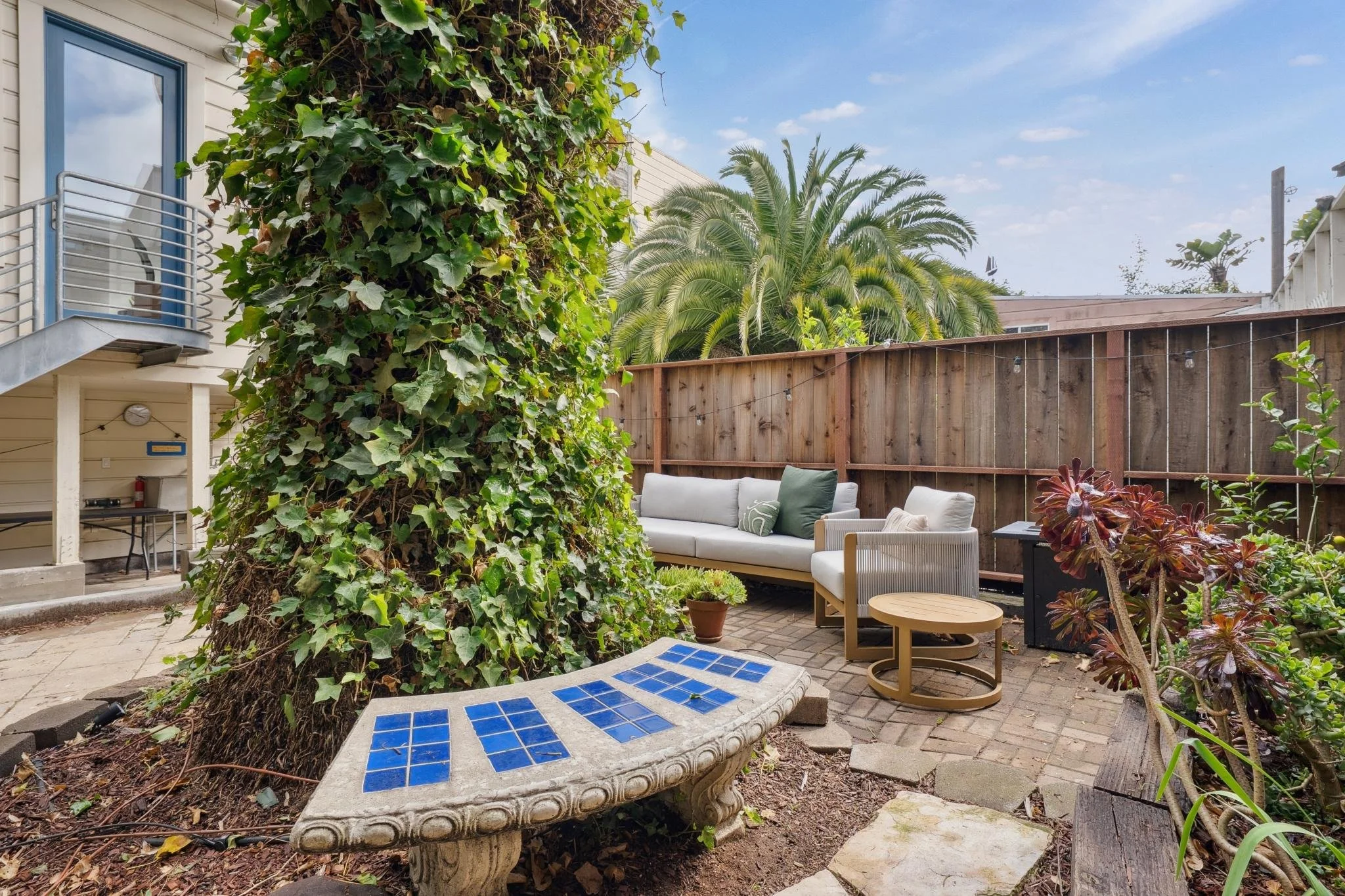 Outdoor backyard patio with a large vine-covered tree, a white outdoor sofa with green and white pillows, a small round table, a decorative bench with blue tiles, a wooden fence, and tropical trees in the background.