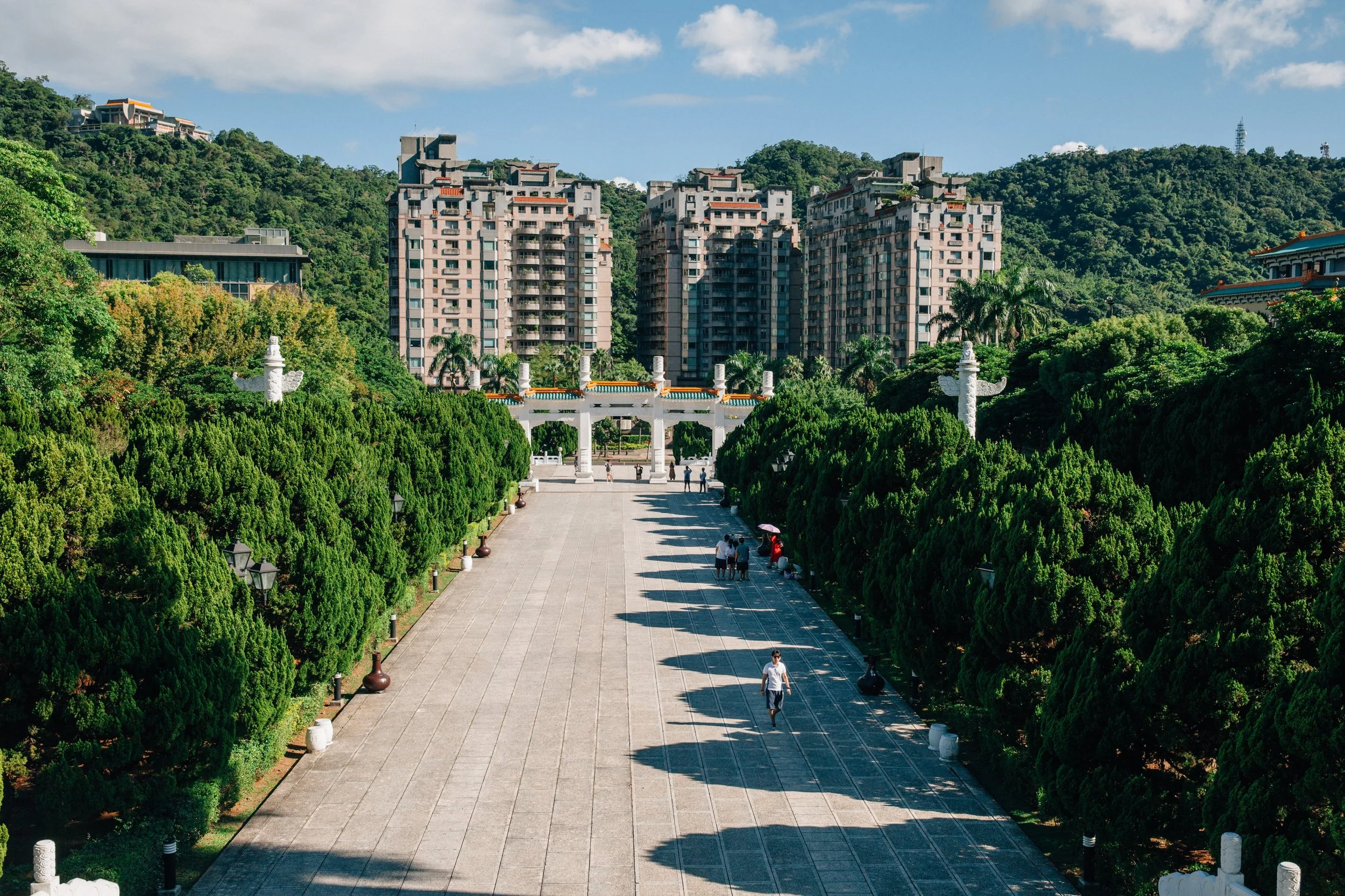 A scenic view of a park with green trees, a wide tiled pathway, and people walking, with residential buildings and mountains in the background.