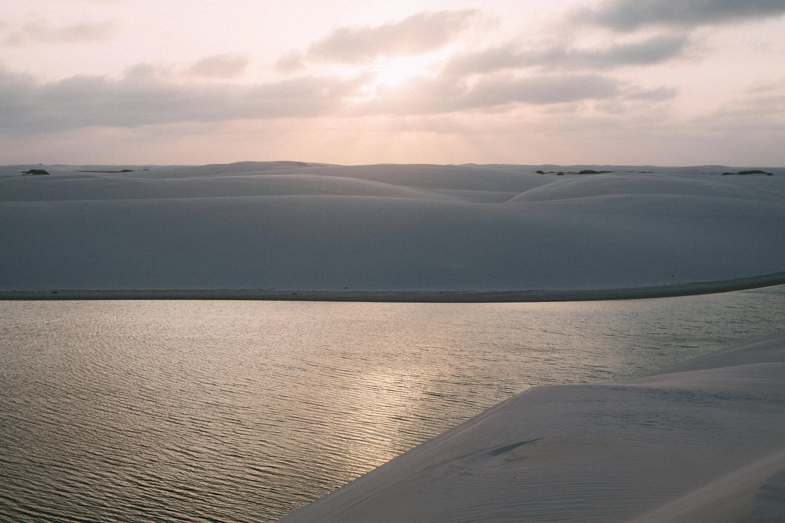 Sand covered landscape with water and a cloudy sky at sunset.