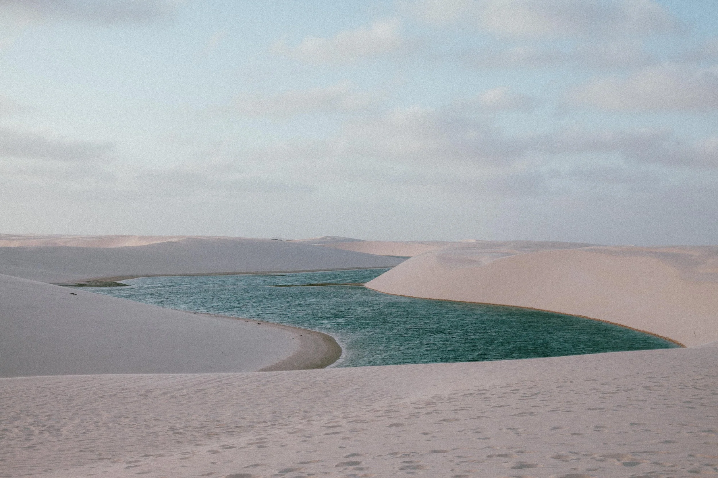 A desert landscape with white sand dunes and a narrow, winding blue-green river flowing through the dunes.