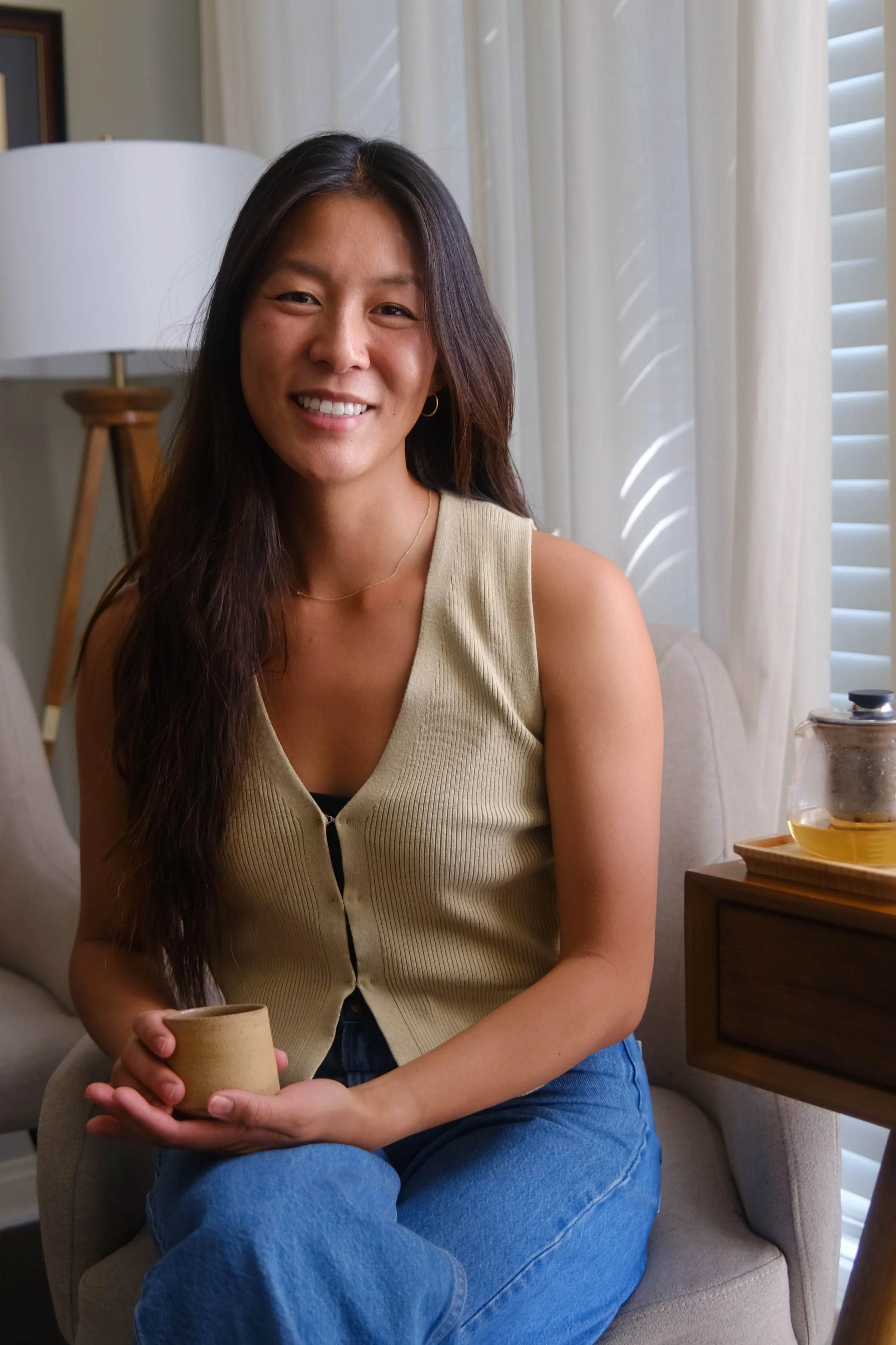 A woman with long dark hair sitting on a beige armchair in a cozy living room, holding a beige coffee mug, smiling at the camera, with a wooden side table and a glass teapot nearby.