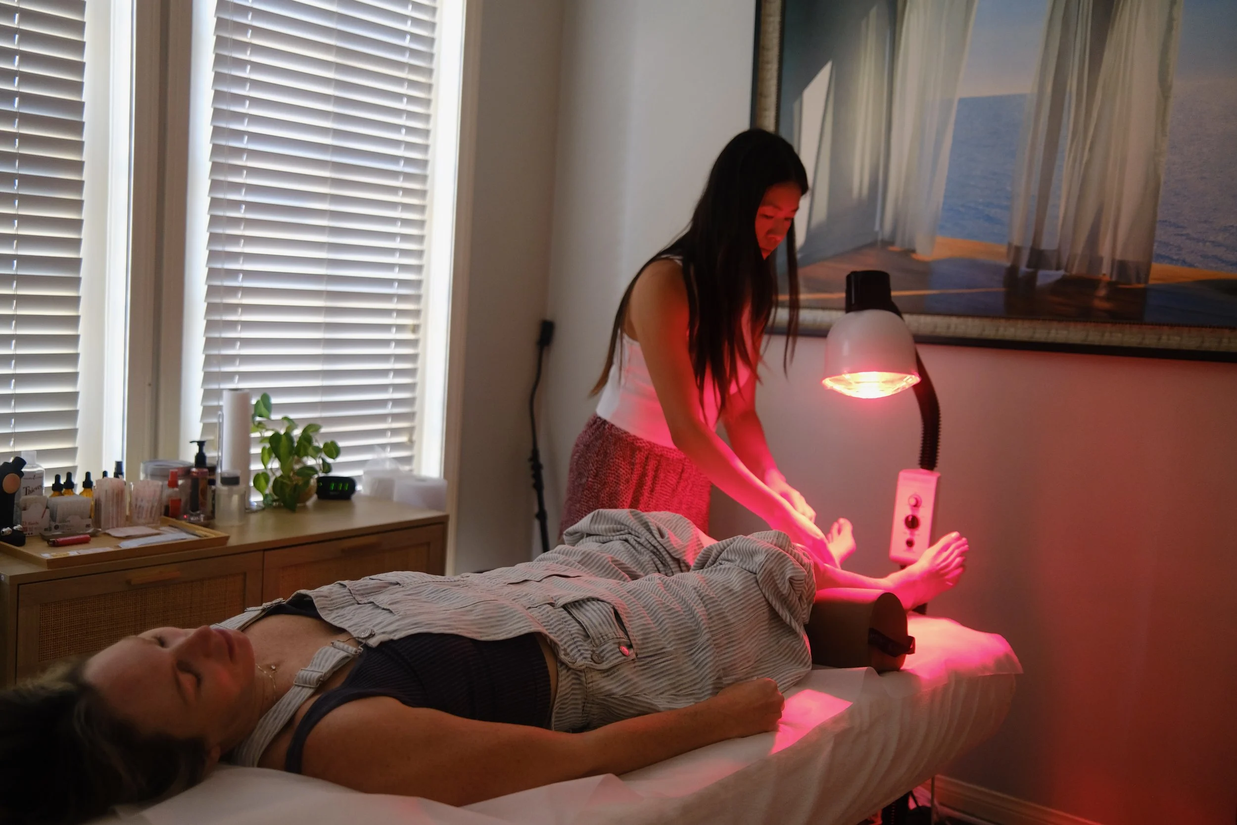 A woman receiving red light therapy treatment on her legs while lying on a bed, with a therapist standing nearby in a room with a window, a painting, and a side table.