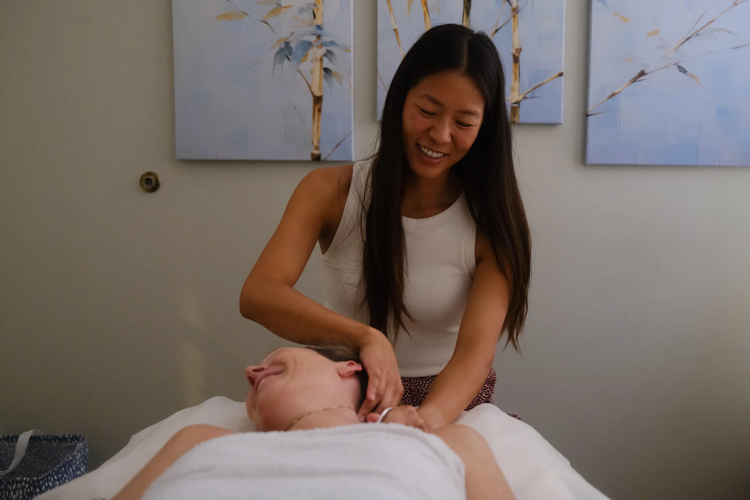 A woman smiling while giving a massage to a person lying on a bed in a room decorated with blue floral paintings.