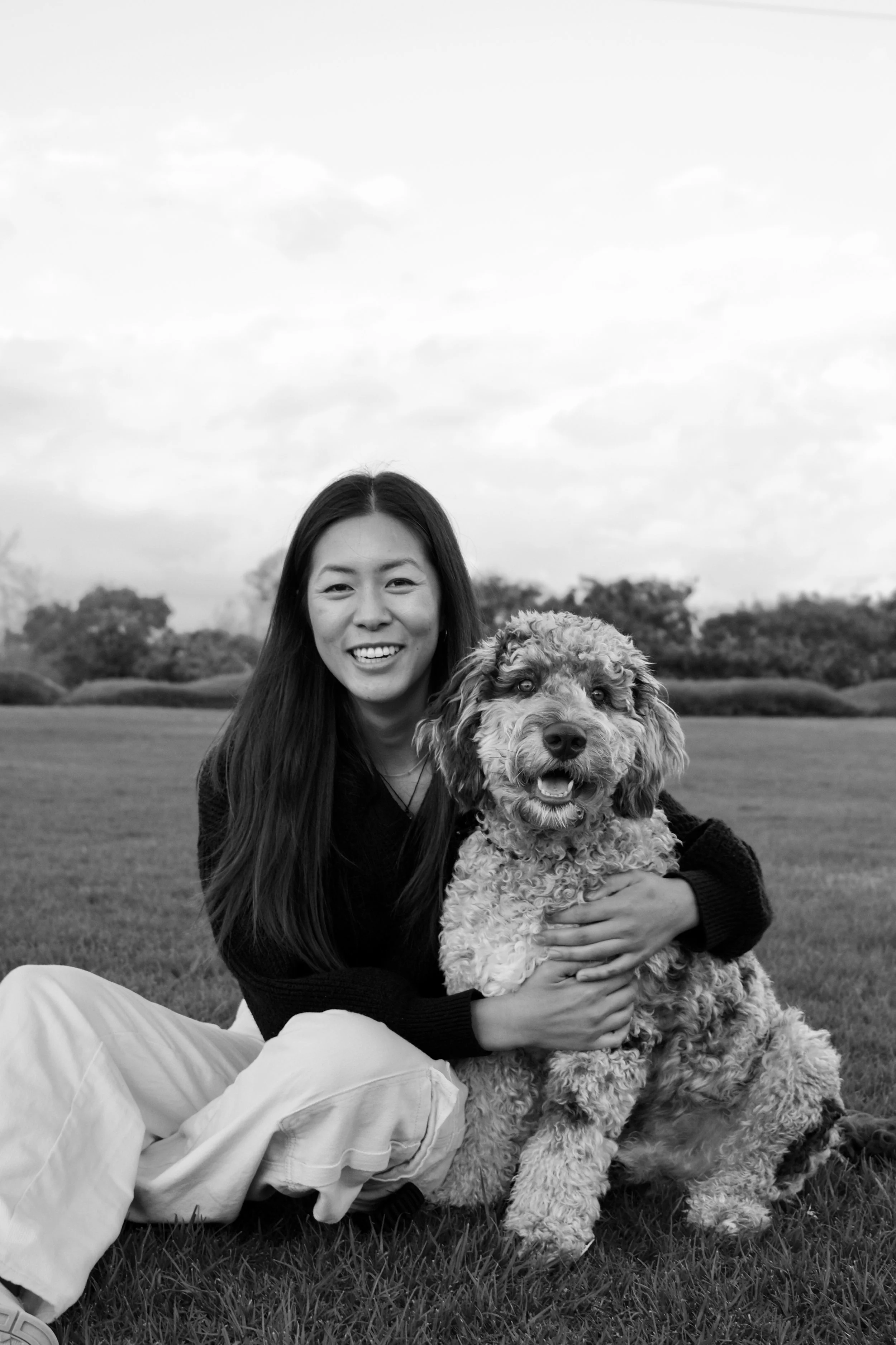 A woman with long hair smiling and sitting on grass, holding a curly-haired dog, outdoors with a cloudy sky and trees in the background.