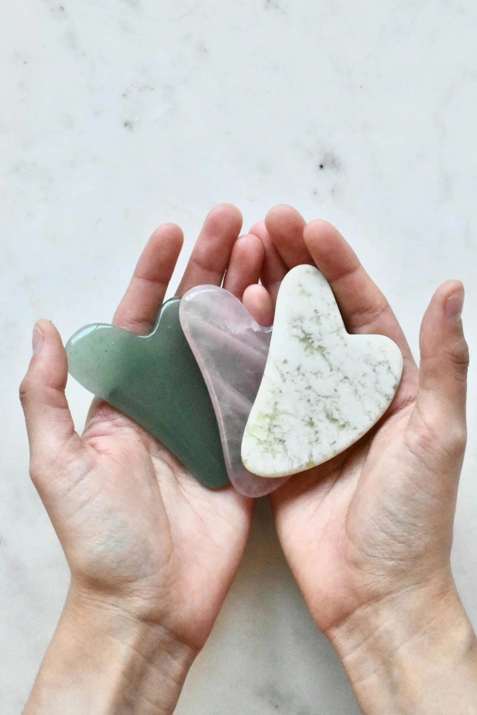 A pair of hands holding three gua-sha stones of green, pink, and white color with marbled patterns.