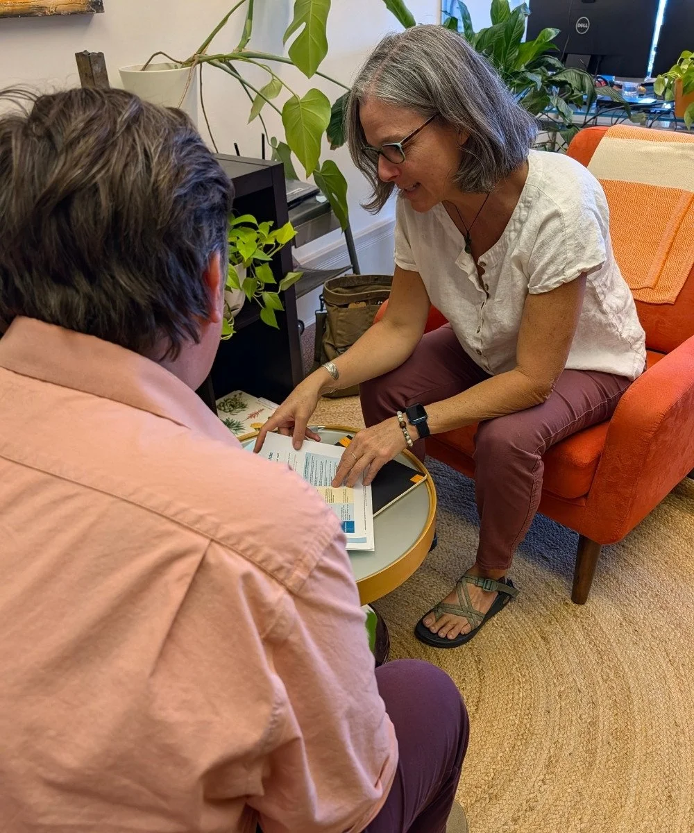 A woman with gray hair and glasses sits on an orange couch, showing a book or magazine to a man with brown hair, who is seated at a small round table. They are in a room with green plants and a computer monitor in the background.