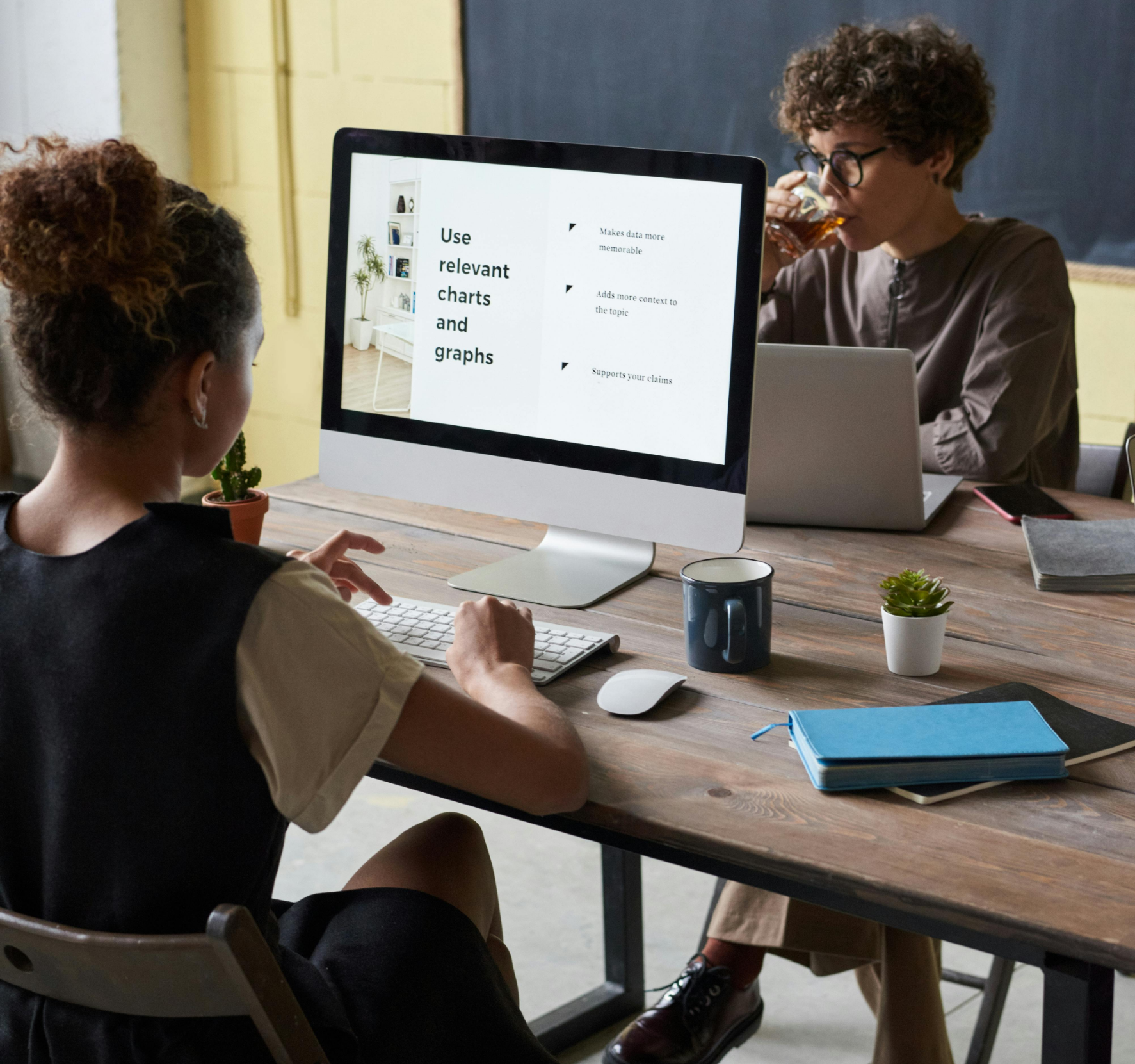 Two people working in an office setting. One person is using a computer displaying text about using relevant charts and graphs. The other person is using a laptop and drinking a beverage. There are potted plants, notebooks, and a mug on the table.