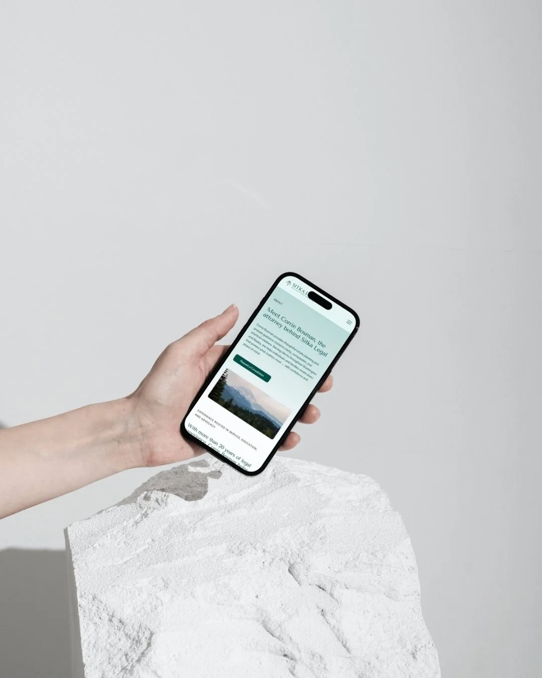 A hand holding a smartphone displaying a website about a lawyer, with a landscape image on the screen, placed on a white textured surface against a plain white background.