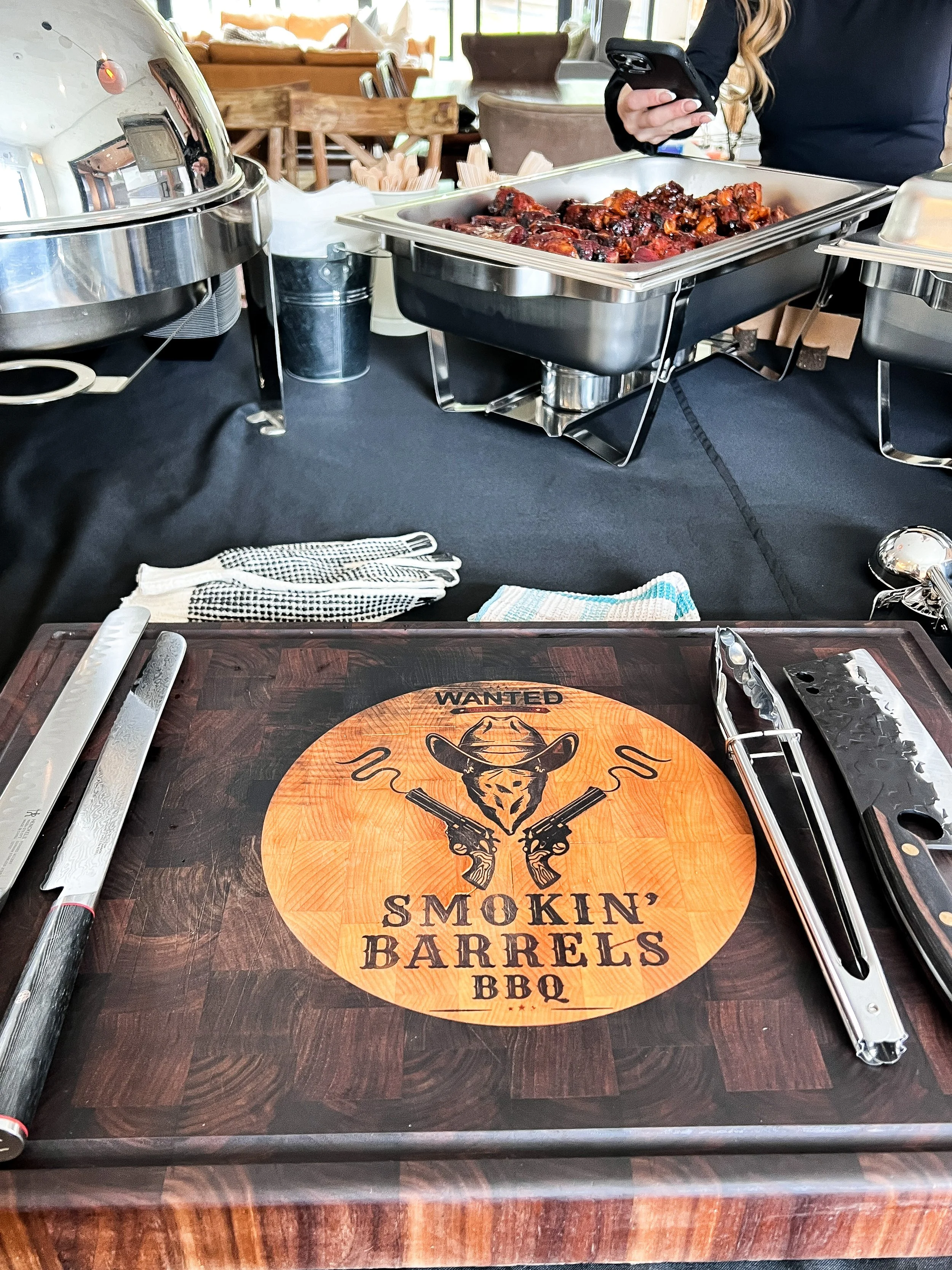Buffet table with trays of barbecue meats, a pair of tongs and a carving knife on a cutting board labeled 'Smokin' Barrels BBQ' with a cowboy hat and guns graphic.