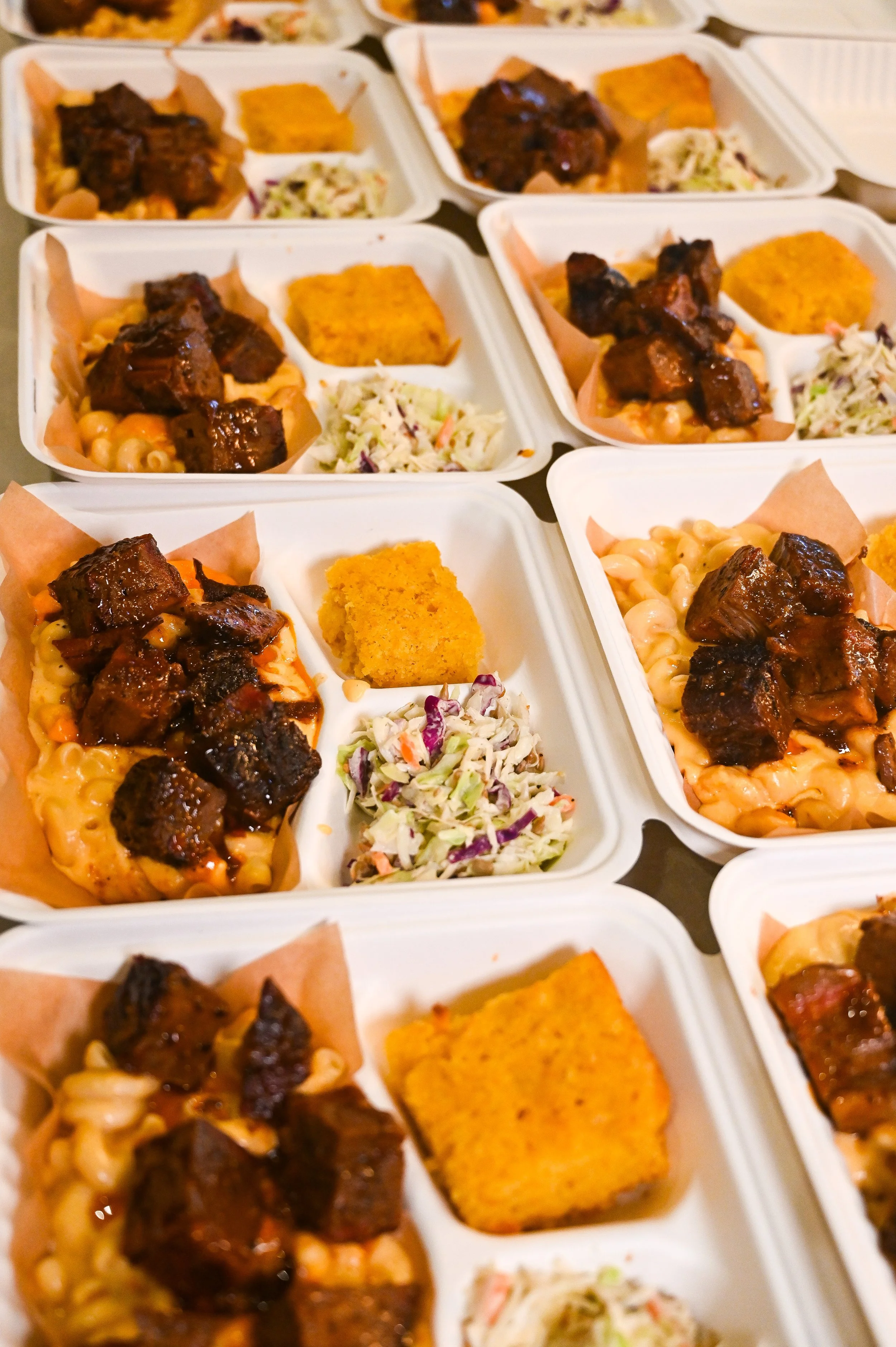 Multiple to-go trays of food containing barbecue beef brisket, macaroni and cheese, coleslaw, and cornbread.