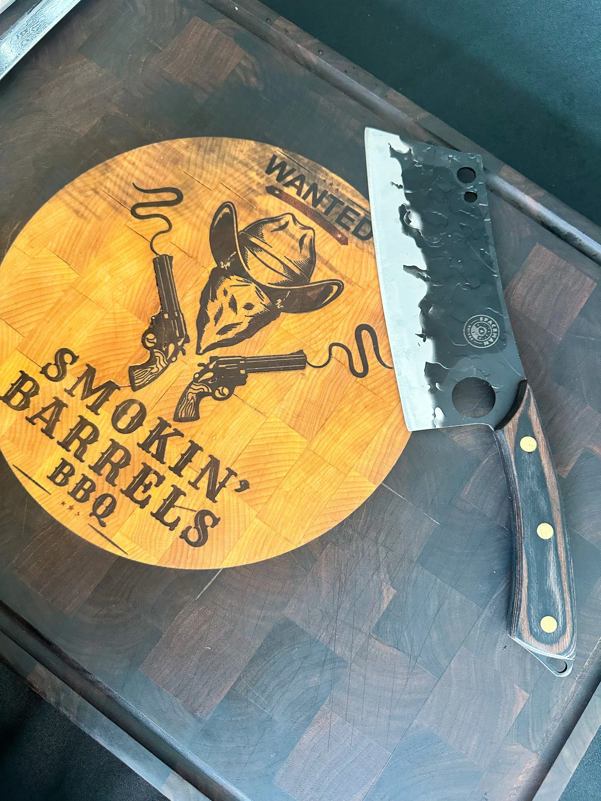 A charcoal grill spatula resting on a wooden table with a circular wood design and a cow illustration. The table has text saying "WANTED, SMOKIN' BARRRELS BBQ".