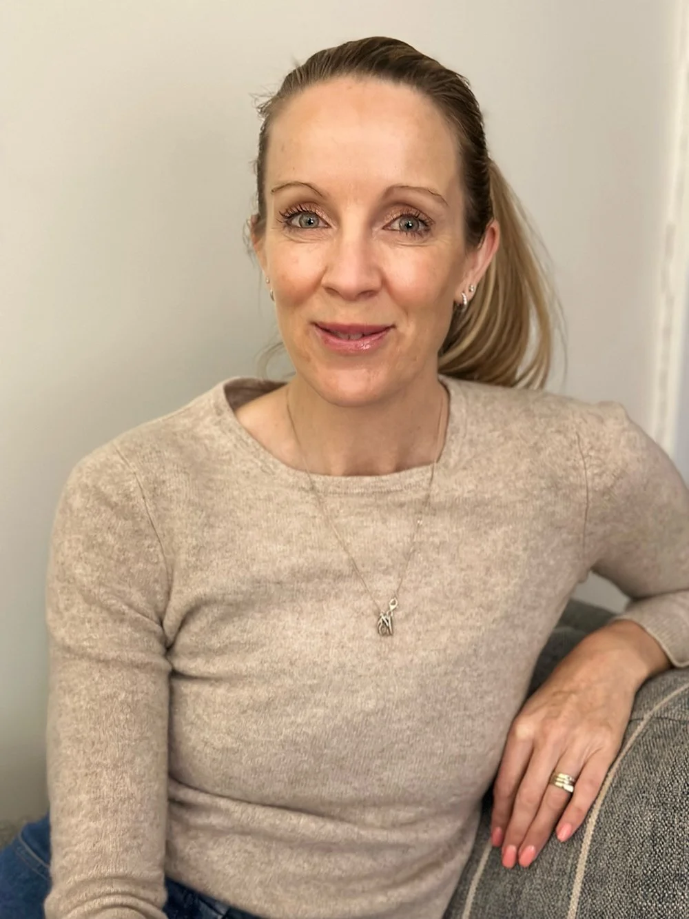 A woman with blonde hair tied back, wearing a beige sweater and jewelry, sitting on a gray couch, smiling at the camera.