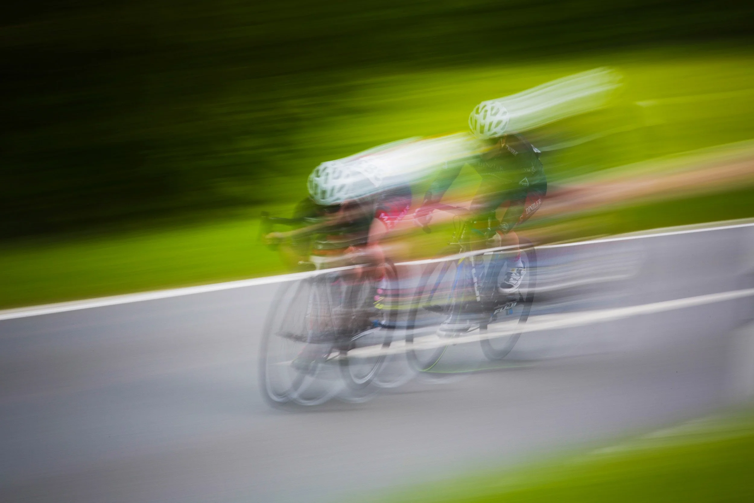 Blurred image of two cyclists racing on a road with green grass in the background.