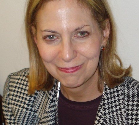 A woman with shoulder-length blond hair, wearing a black and white houndstooth patterned blazer and a black top, smiling at the camera.