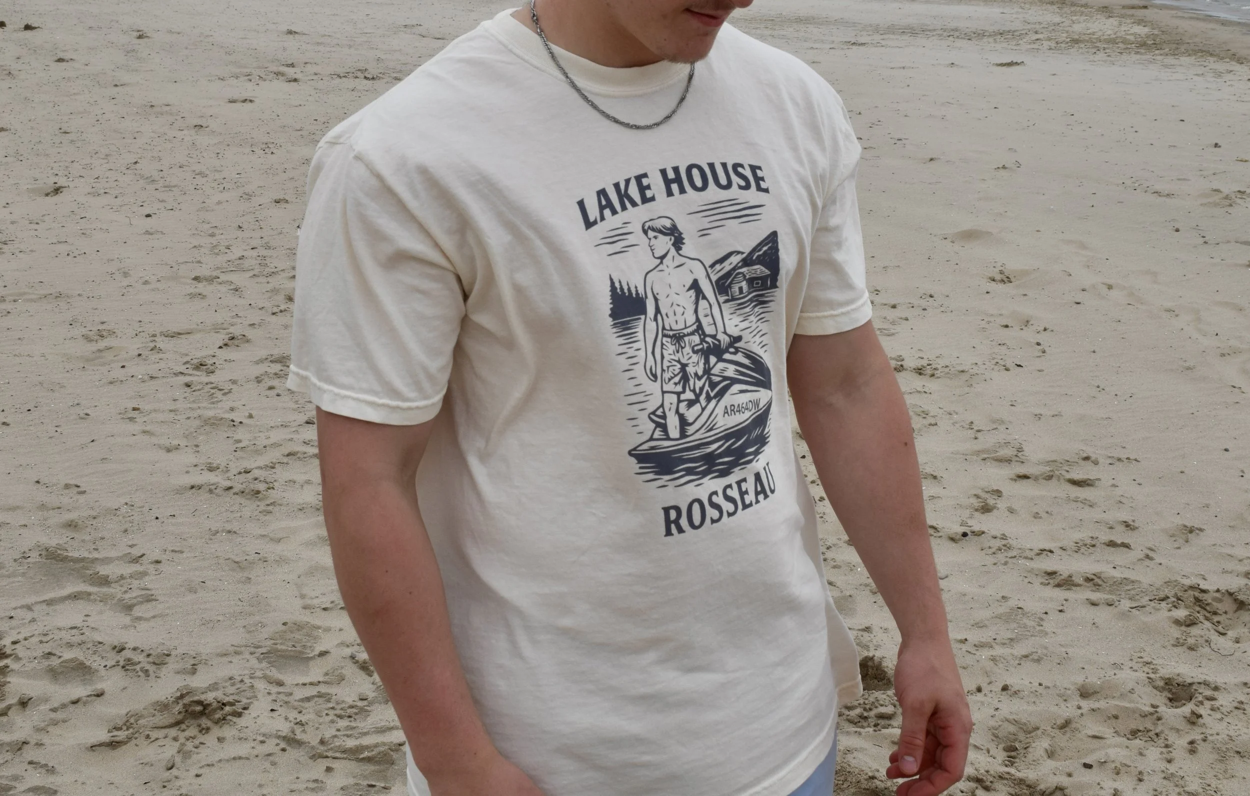 Person standing on a sandy beach wearing a light-colored T-shirt with a graphic design and text Reading 'Lake House Rosseau'.