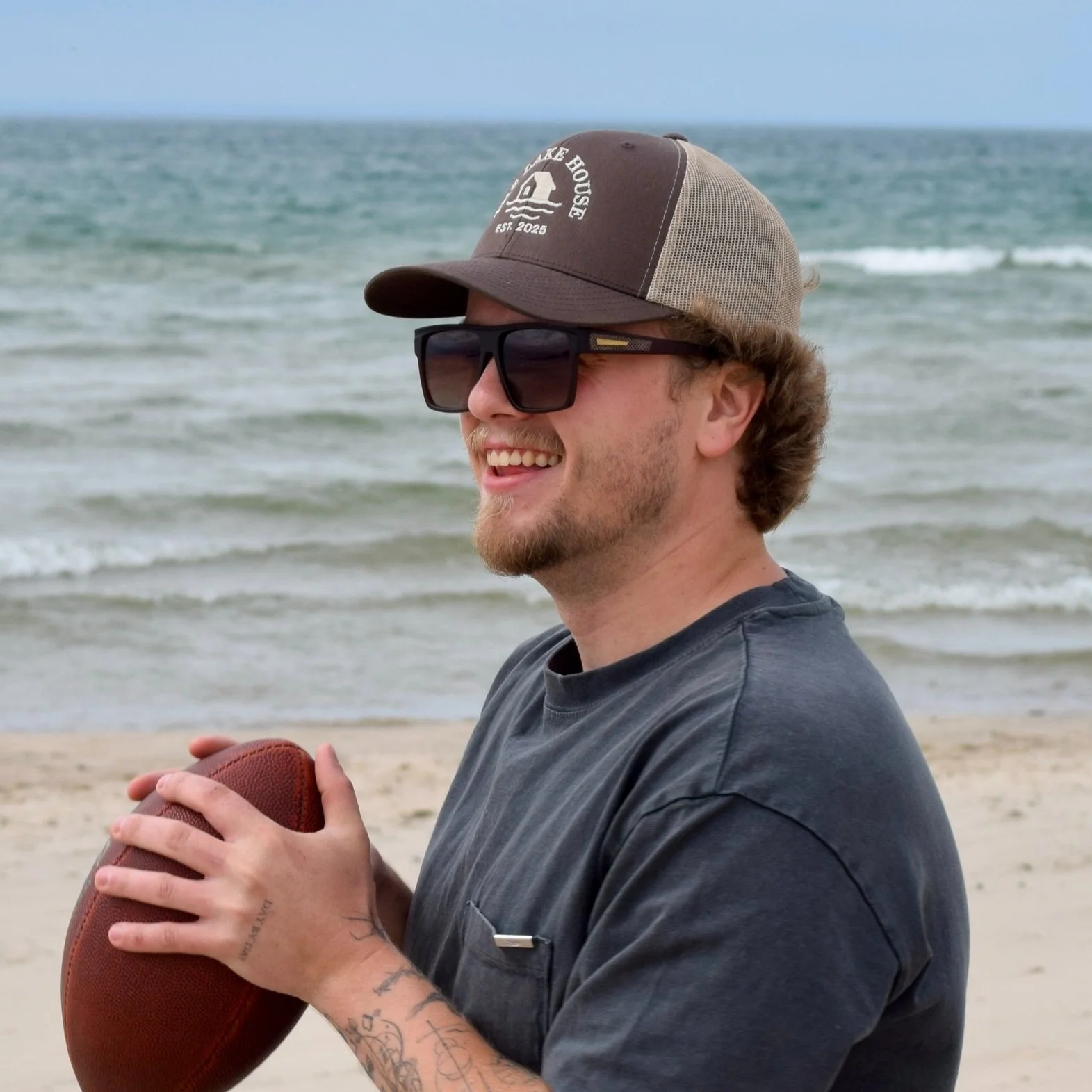 A young man with tattoos on his arm, wearing sunglasses, a cap, and a gray T-shirt, holding a football on a beach with the ocean in the background.
