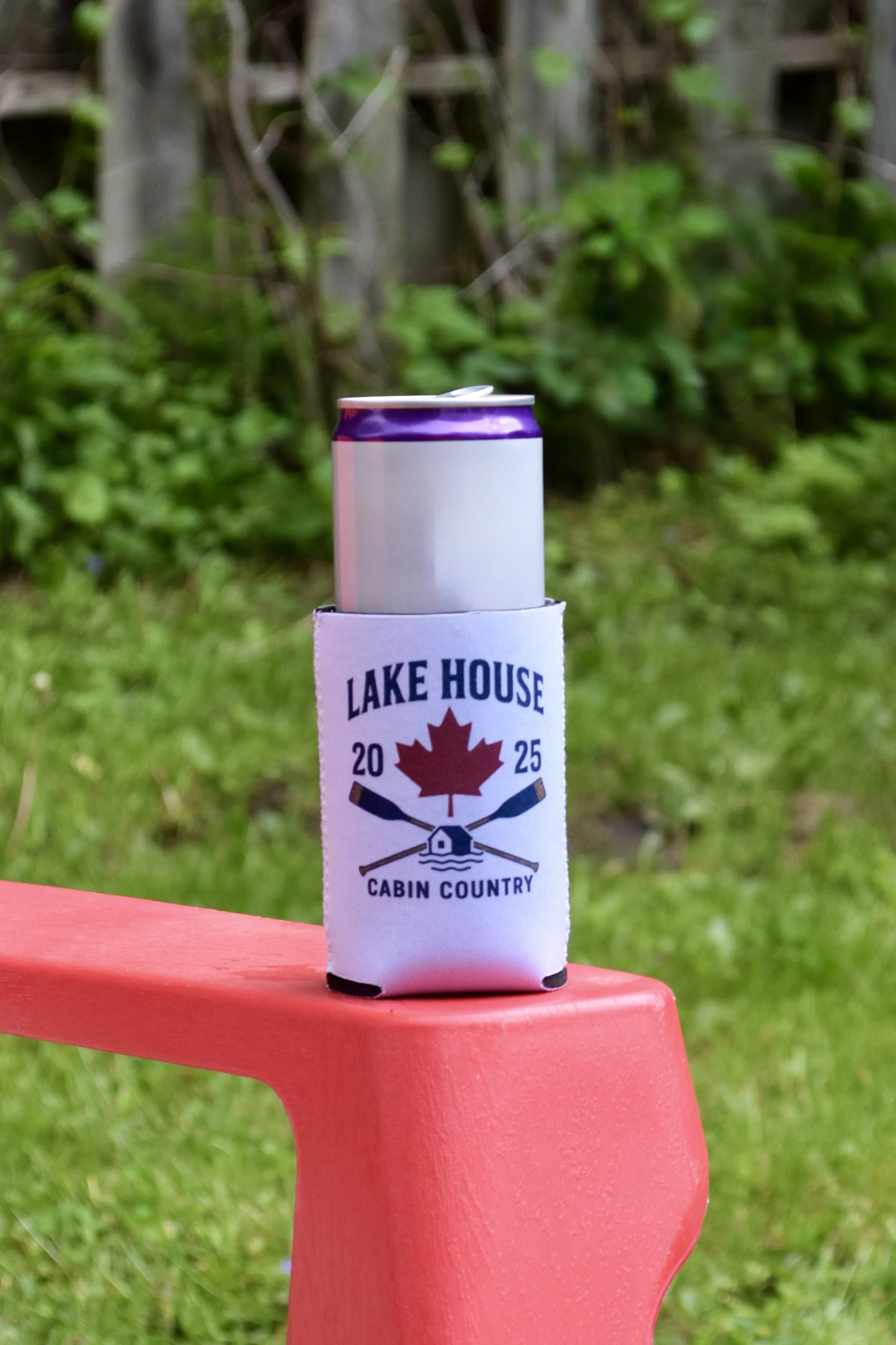 A soda can in a foam koozie with a logo that says 'Lake House Cabin Country' and a red maple leaf, on a pink plastic chair outdoors with a grassy yard and trees in the background.