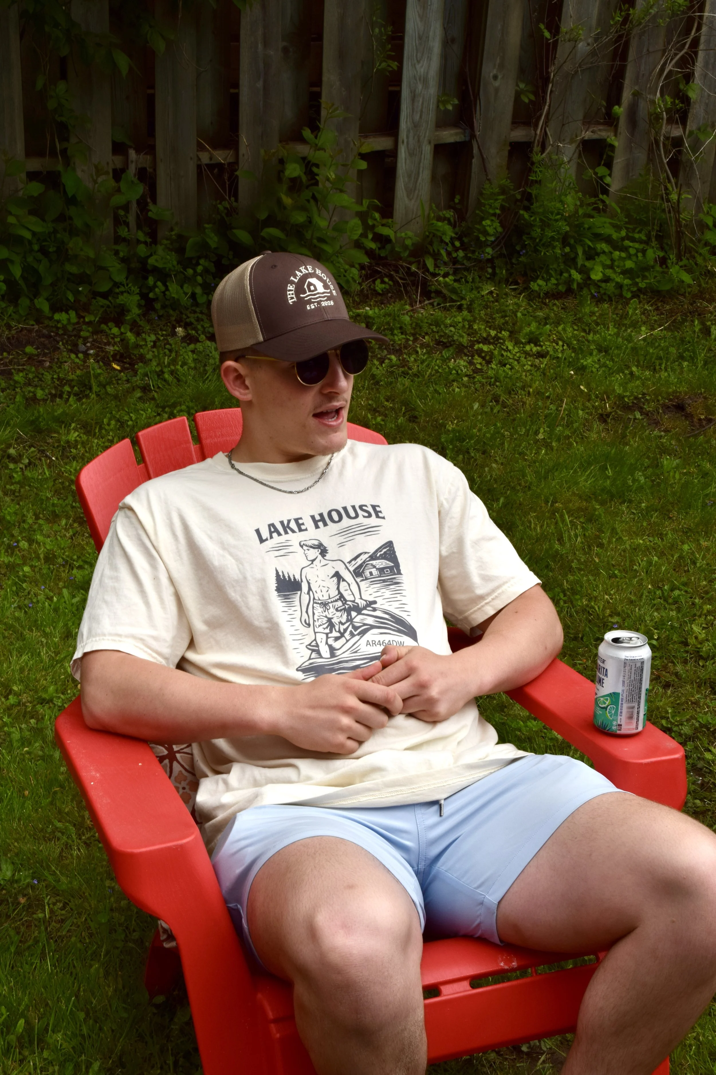 Young man sitting in a red outdoor chair, wearing sunglasses, a baseball cap, a t-shirt, and shorts, with a soda can on the armrest, in a backyard with grass and a wooden fence.