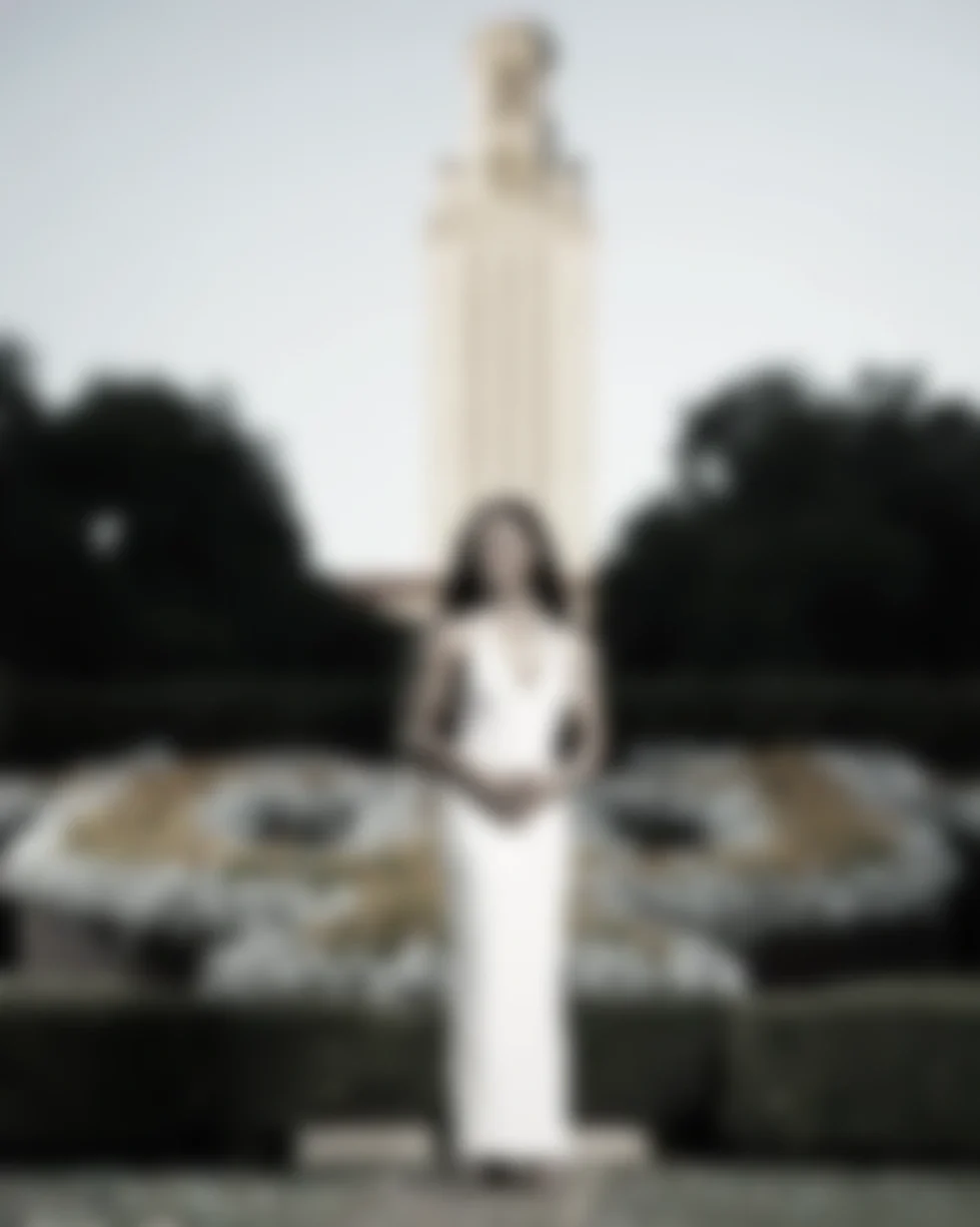 A woman standing in front of a wedding cake with two or three tiers, decorated with gold accents and topped with what appears to be white flowers, in an outdoor setting with trees and a tall building in the background.
