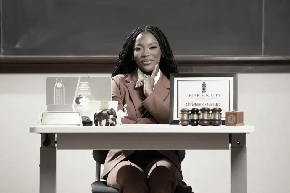 A woman sitting at a desk in a classroom, with a blackboard behind her. The desk has various items, including framed certificates, jars, small statues, and writing supplies.