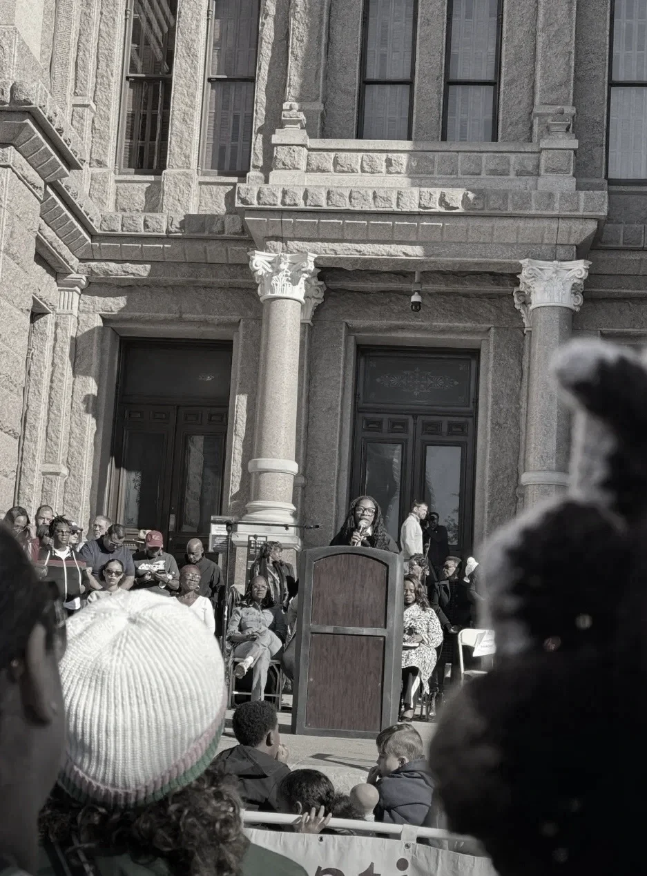 A woman speaking at a podium during a public gathering outside a historic stone building with columns.