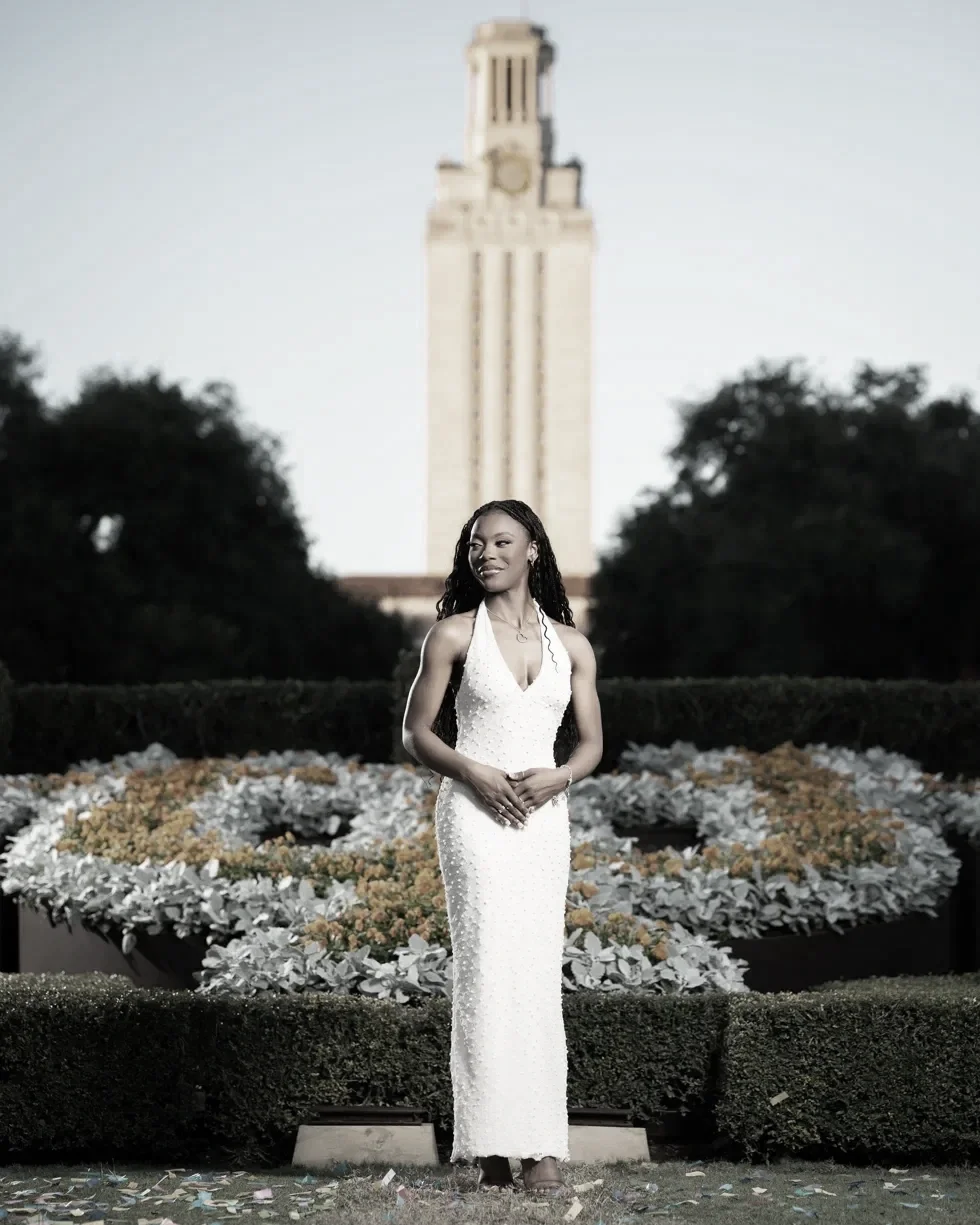 A woman in a white dress standing outdoors on a grassy area in front of a flowerbed and trees, with a tall building in the background.