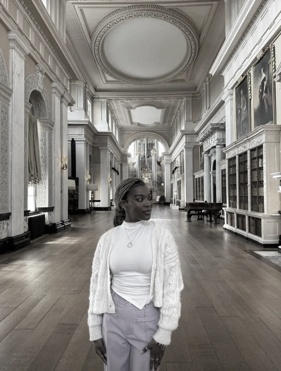 A woman in a white turtleneck, cardigan, and light-colored pants stands in a grand, historic library with ornate ceiling details, tall bookshelves, and a large organ at the far end.