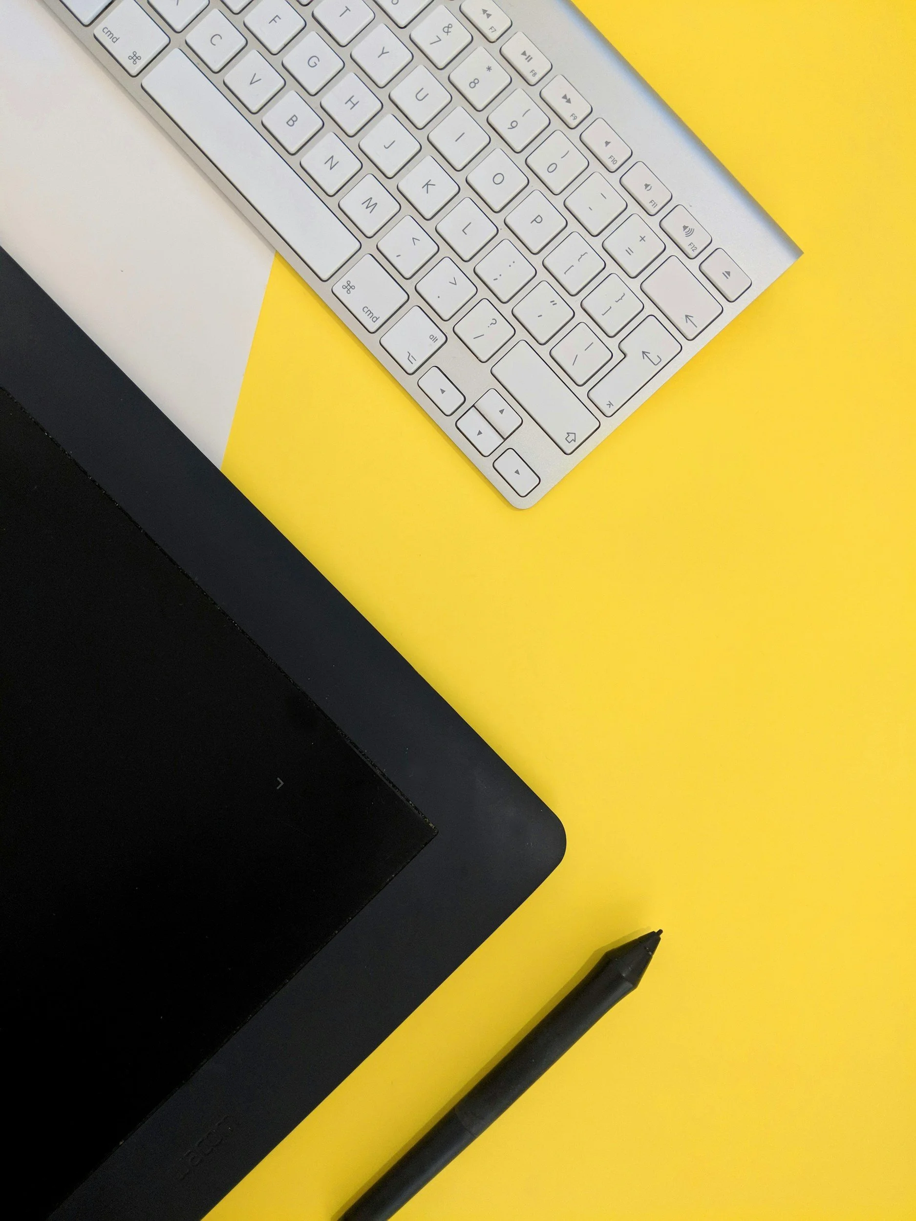 A top view of a workspace with a black drawing tablet, a black stylus pen, a white keyboard, and a yellow surface in the background.