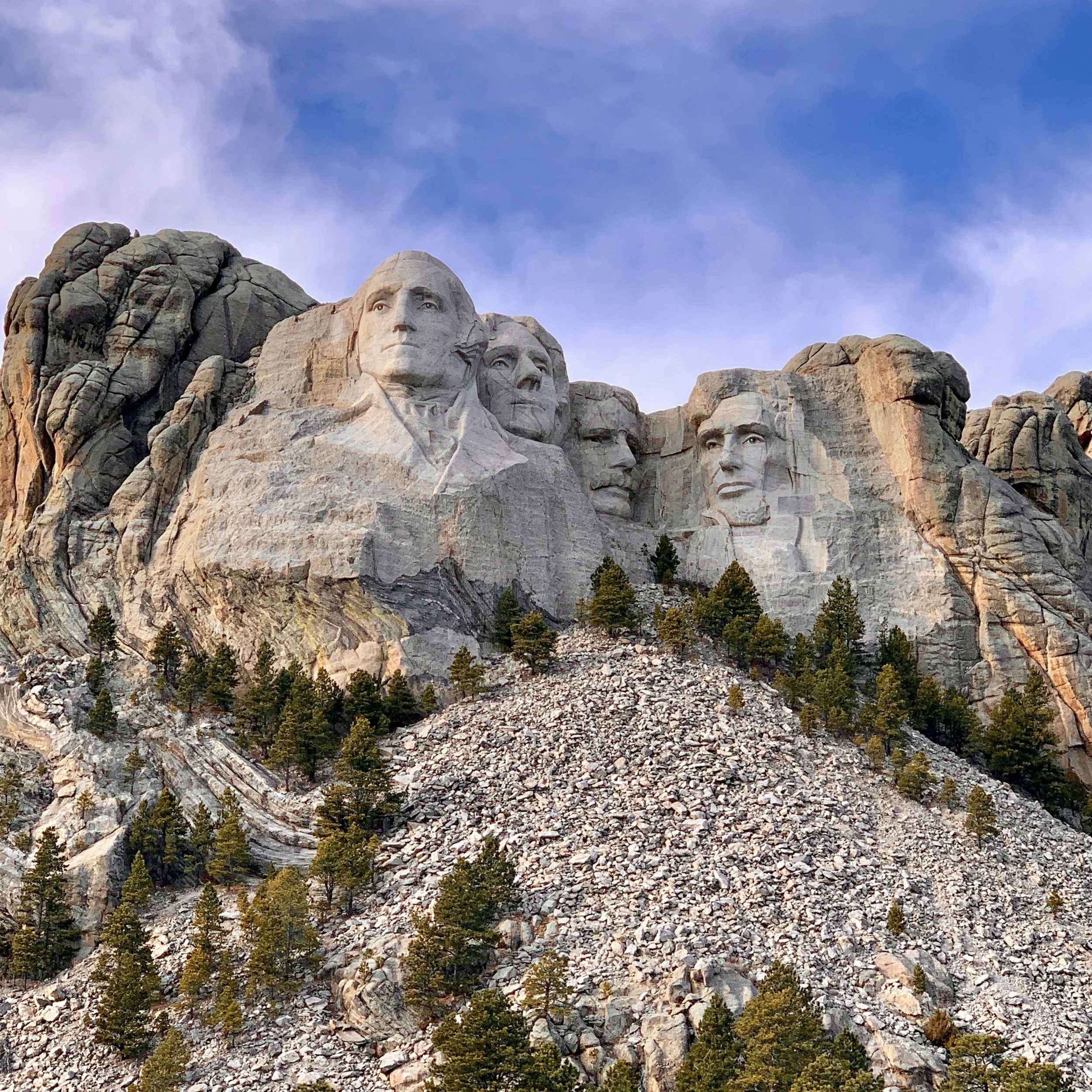 Close-up photograph of Mount Rushmore, featuring the carved faces of four U.S. presidents, set against a partly cloudy sky and surrounded by pine trees and rocky terrain.