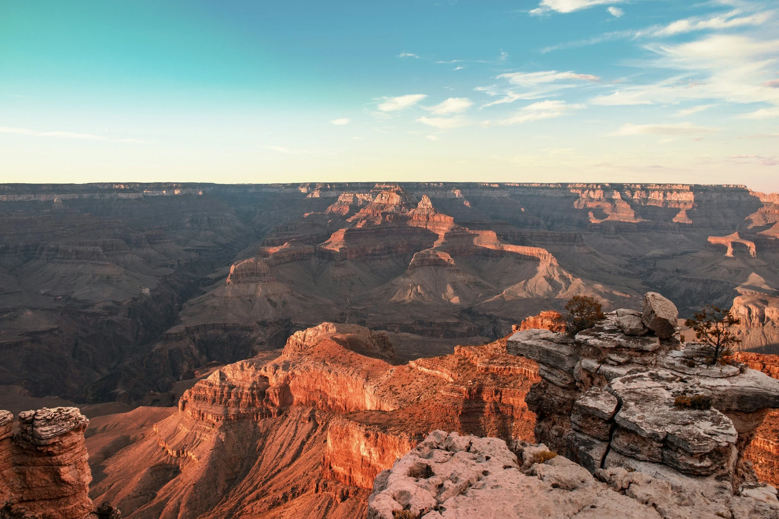 A view of the Grand Canyon during sunset with orange and red rock formations, a rocky foreground, and a partly cloudy sky.