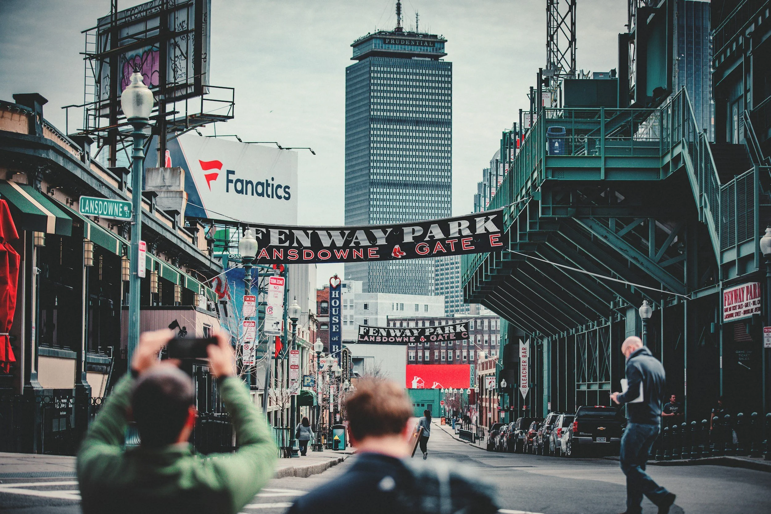 View of a street scene in Fenway Park, Boston, featuring signs, pedestrians, and a tall building in the background, with some people taking photos.