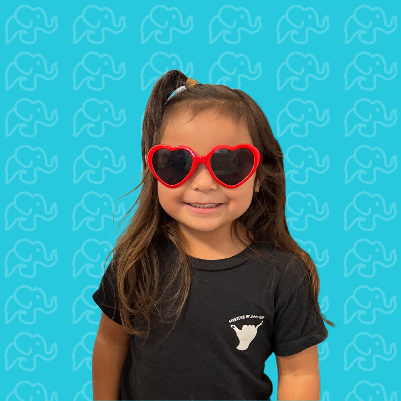 A young girl wearing heart-shaped sunglasses and a black t-shirt smiling in front of a blue background with a repeating elephant pattern.