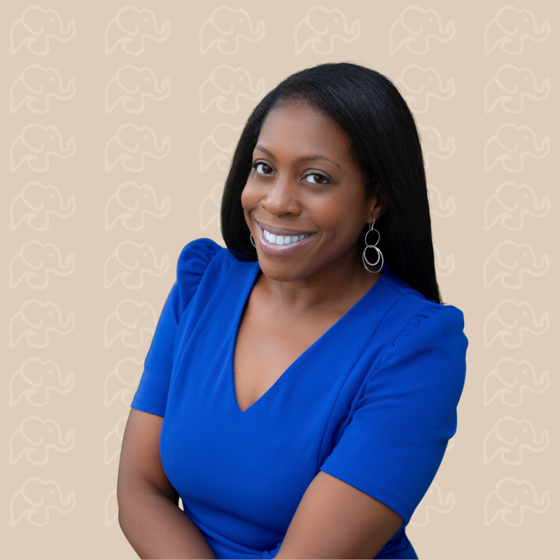 A woman with dark hair and a bright smile wearing a royal blue top and silver hoop earrings, standing against a beige background with a subtle elephant pattern.