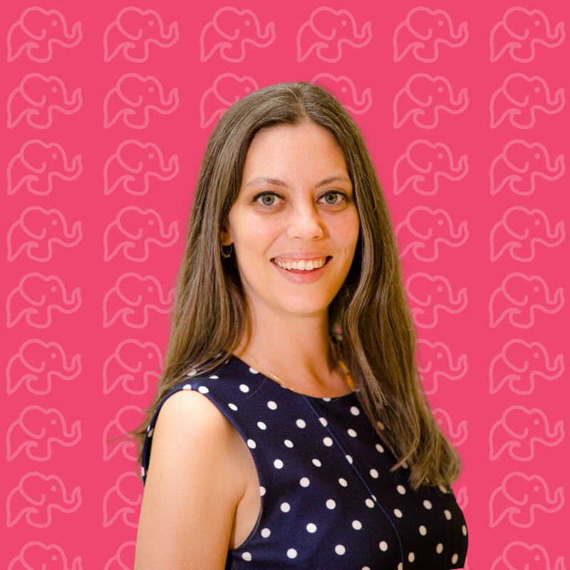 Portrait of a woman with long brown hair, wearing a navy blue dress with white polka dots, smiling against a pink background with a repeating elephant outline pattern.