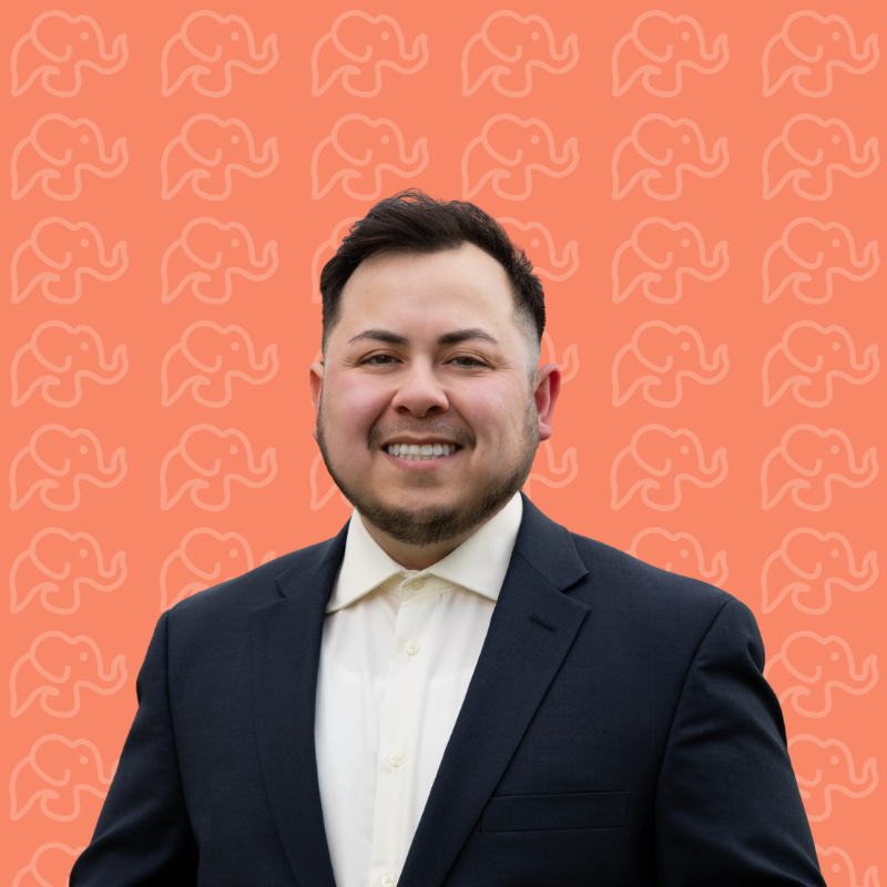 Headshot of a man wearing a dark suit and white shirt smiling against an orange background with a subtle pattern of elephants.