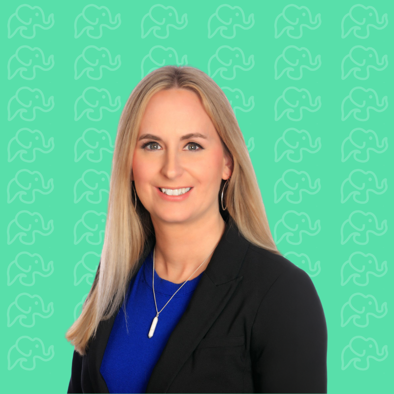 Professional woman with long blonde hair, wearing a black blazer and blue top, smiling against a green background with elephant pattern.