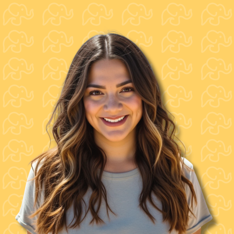 A young woman with long, wavy brown hair, smiling, wearing a light gray t-shirt, standing in front of a yellow background with a subtle pattern of white elephants.