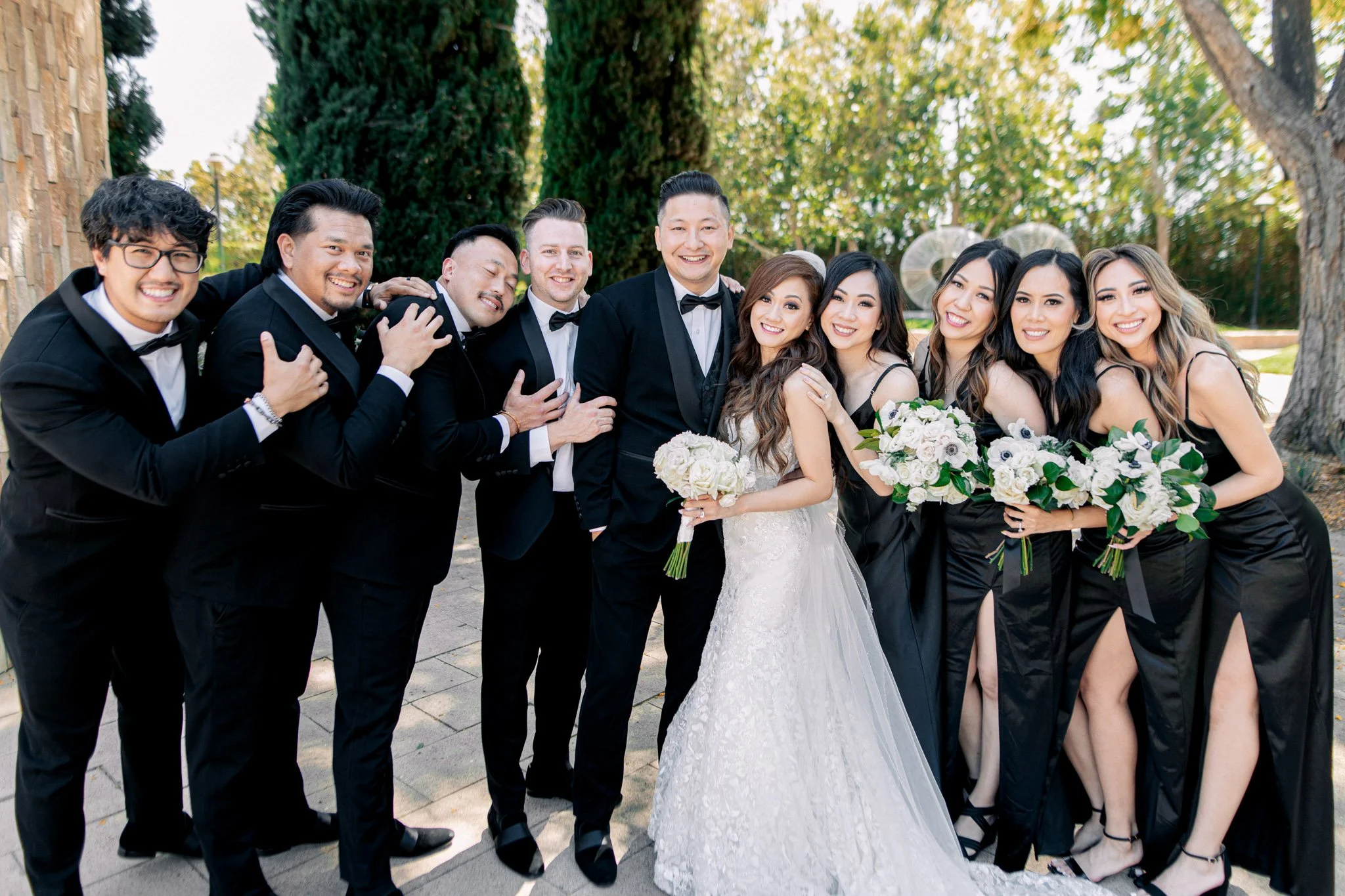 A wedding party with the bride and groom surrounded by friends, all dressed in formal attire, outdoors on a sunny day with trees in the background.